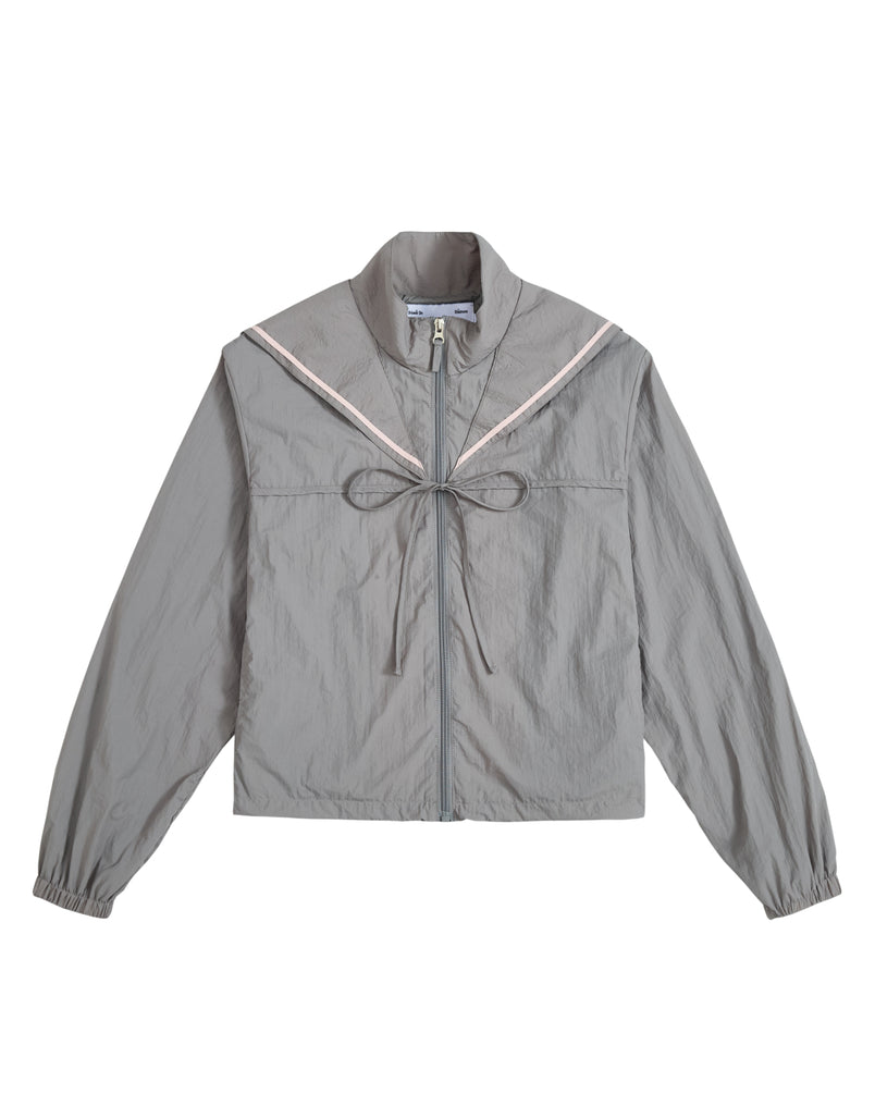 The GREY SAILOR WINDBREAKER by Sundae School is a light gray cropped jacket featuring a high collar, long sleeves, and a decorative tie at the chest. A subtle pink chevron detail on the front evokes the look of a sailor uniform, while the slightly crinkled fabric adds both style and comfort.