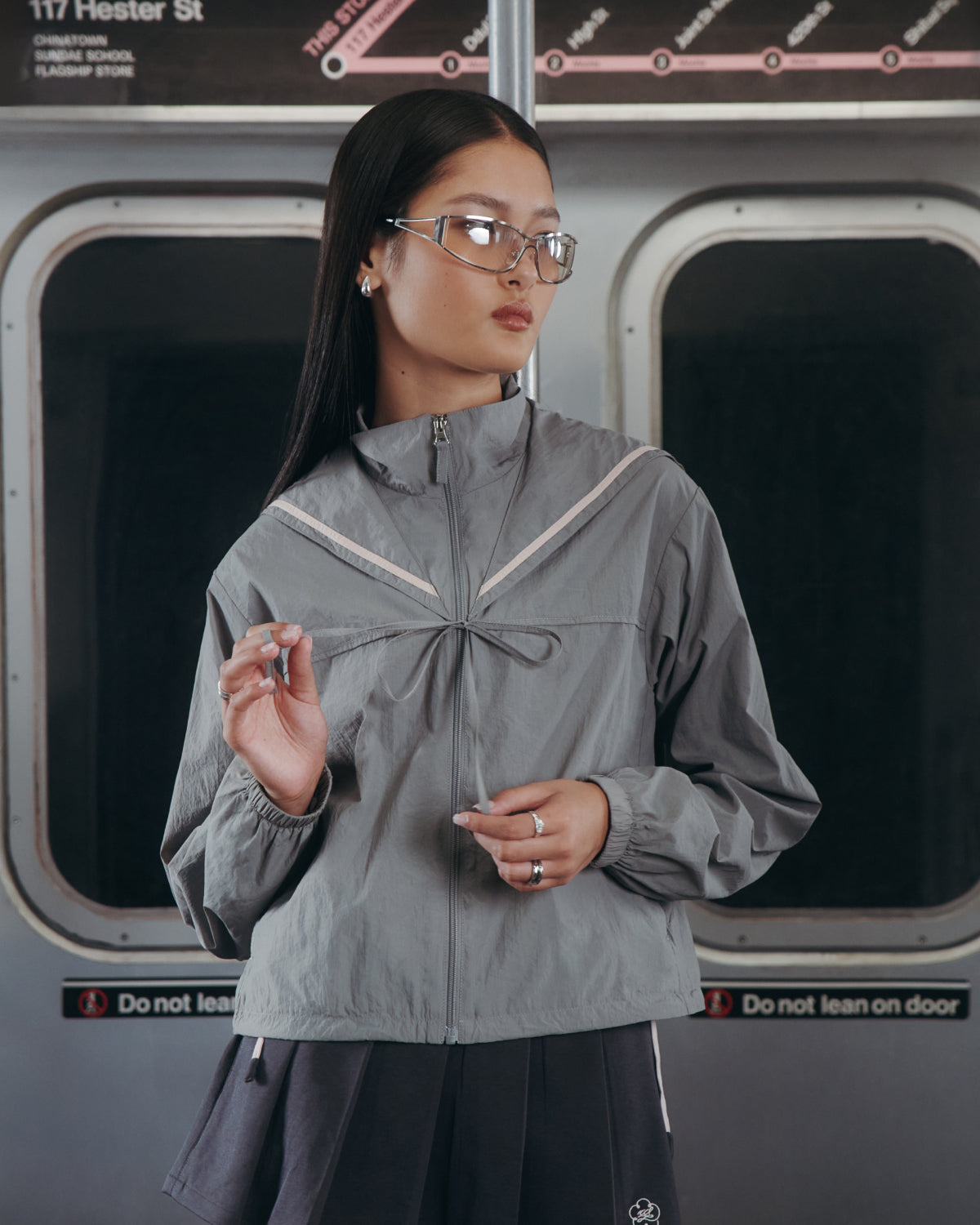 A woman in glasses and the Sundae School GREY SAILOR WINDBREAKER stands in a subway car, holding a pen. Behind her are subway doors and a blurred map as she looks to the side with a calm expression.