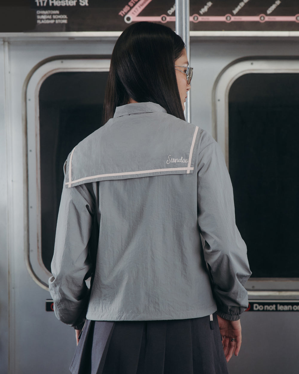 A person with long dark hair, wearing glasses and the GREY SAILOR WINDBREAKER from Sundae School, stands inside a subway train facing away from the camera. Subway signage and windows are visible in the background.