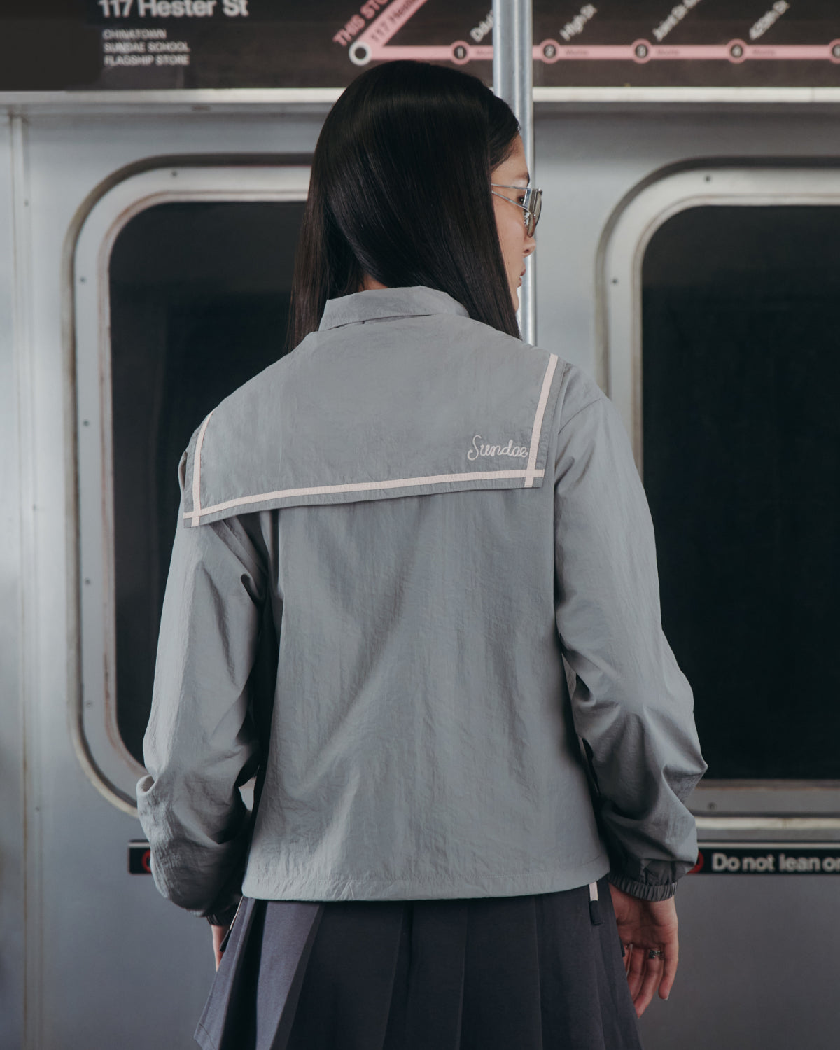 A person with long dark hair, wearing glasses and the GREY SAILOR WINDBREAKER from Sundae School, stands inside a subway train facing away from the camera. Subway signage and windows are visible in the background.
