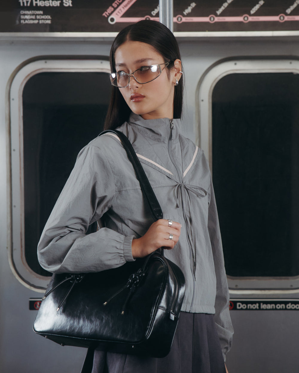 Wearing the GREY SAILOR WINDBREAKER by Sundae School, a person with clear glasses stands in a subway car holding a black handbag. Behind them, subway map details, windows, and signage inside the train are visible.