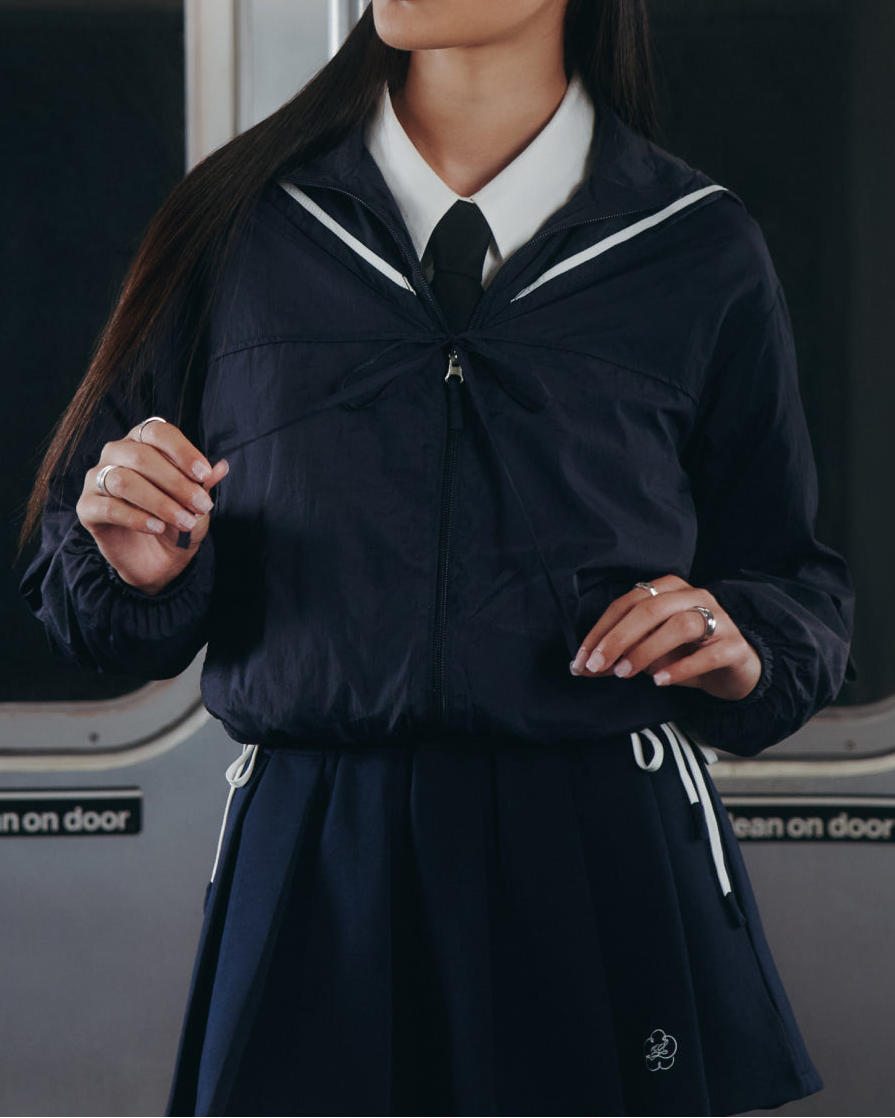 A person in Sundae School's NAVY SAILOR WINDBREAKER and NAVY PLEATED JERSEY MINI SKIRT stands in a subway car.