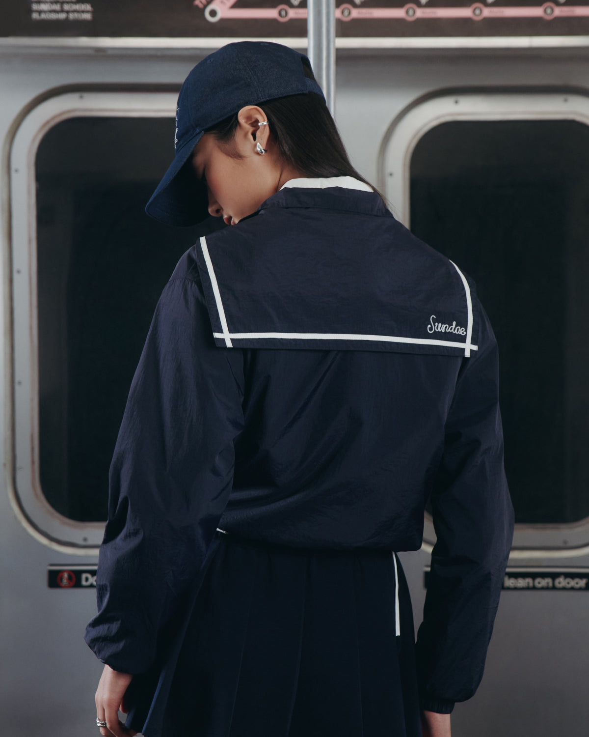 A person in a Sundae School NAVY SAILOR WINDBREAKER and cap stands with their back to the camera on a subway train, 