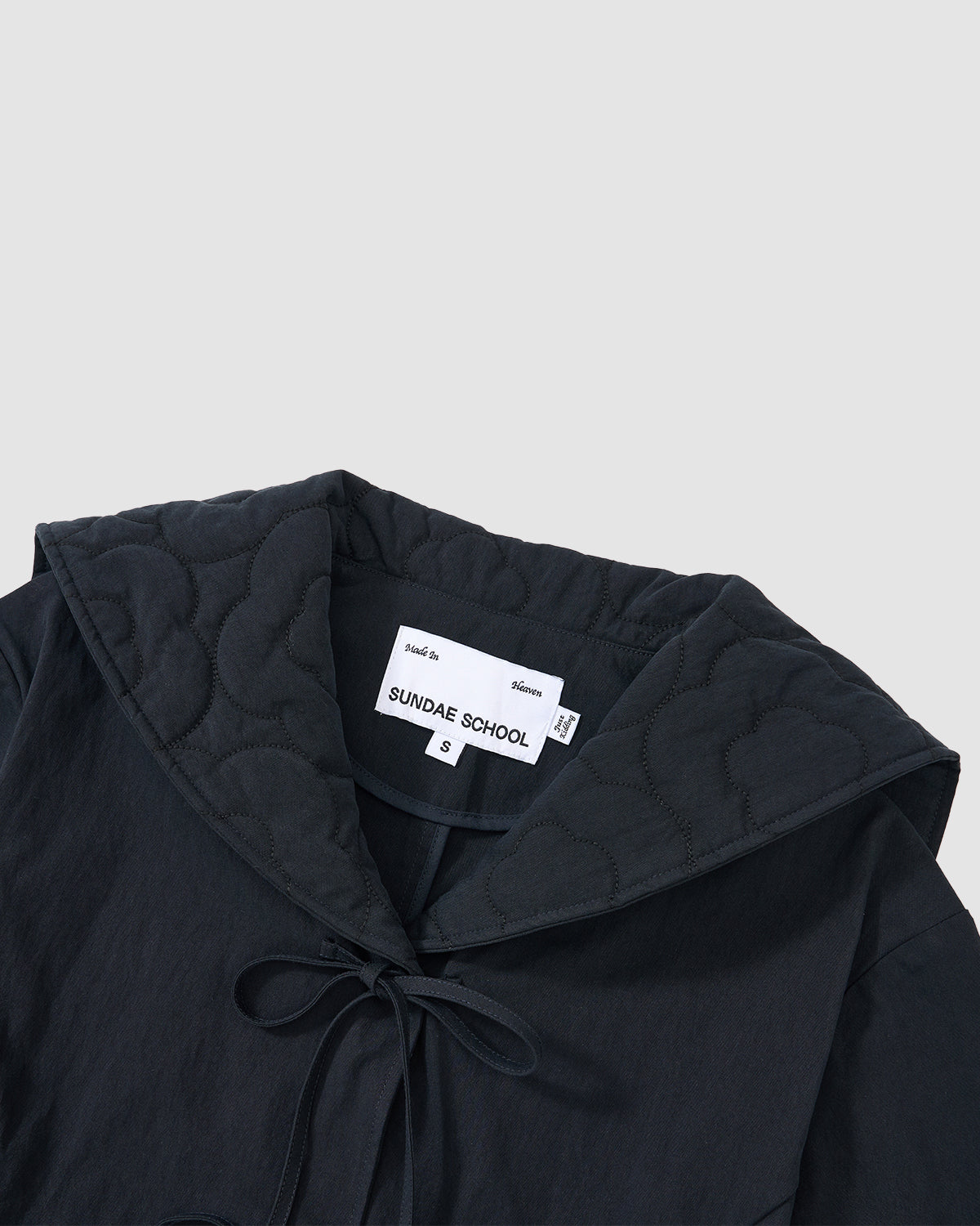 A close-up of the NAVY SAILOR COLLAR UNIFORM JACKET by Sundae School shows mochi quilted stitching, a scalloped-edge collar, and a front ribbon tie. The inner label displays the brand and size S. The background is light gray.