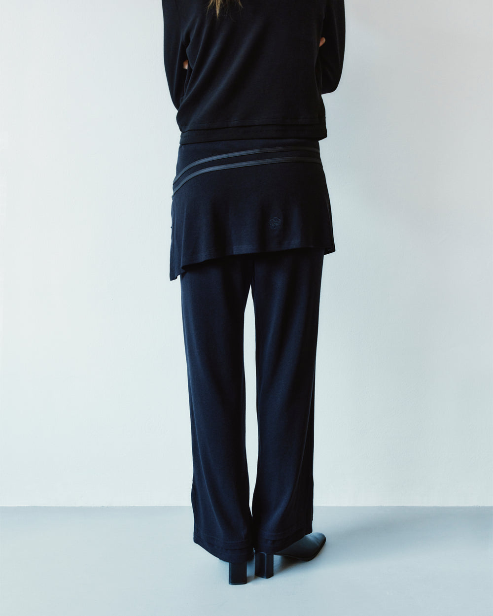 Back view of a person wearing BLACK SEONGSU APRON PANTS, which are black wide-leg trousers featuring an integrated hybrid wrap skirt detail at the waist.