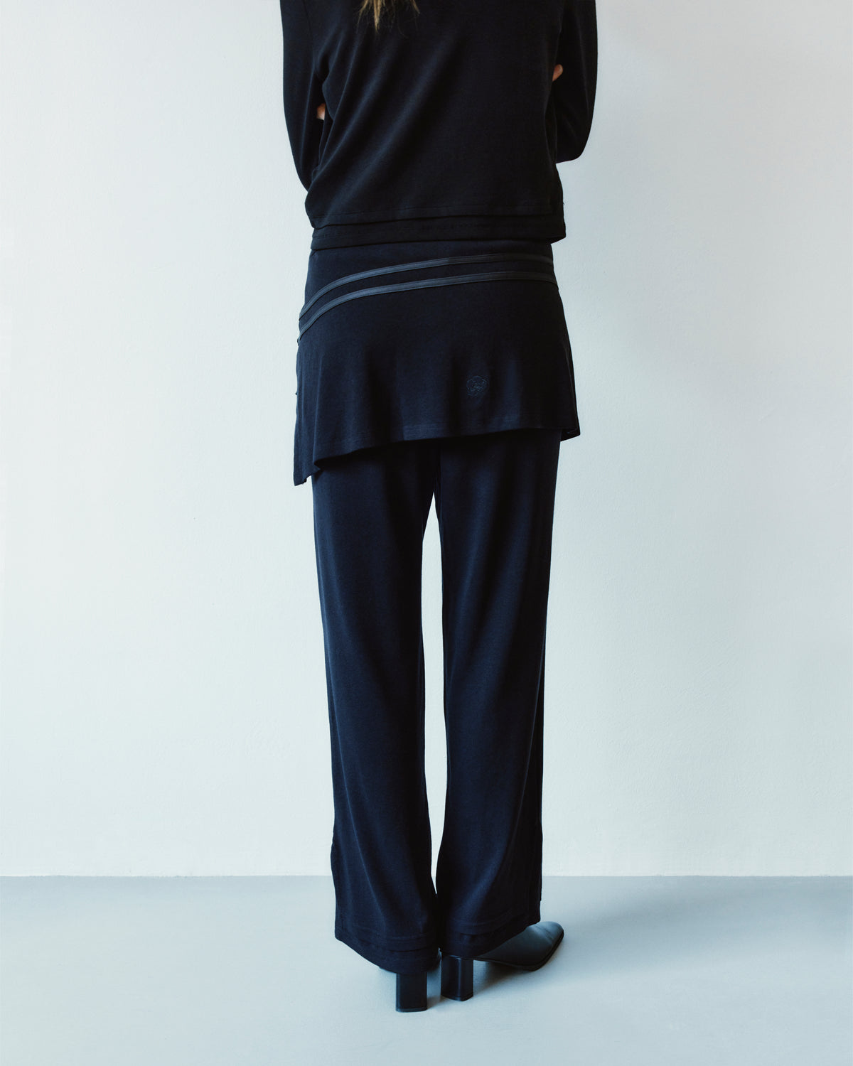 Back view of a person wearing BLACK SEONGSU APRON PANTS, which are black wide-leg trousers featuring an integrated hybrid wrap skirt detail at the waist.