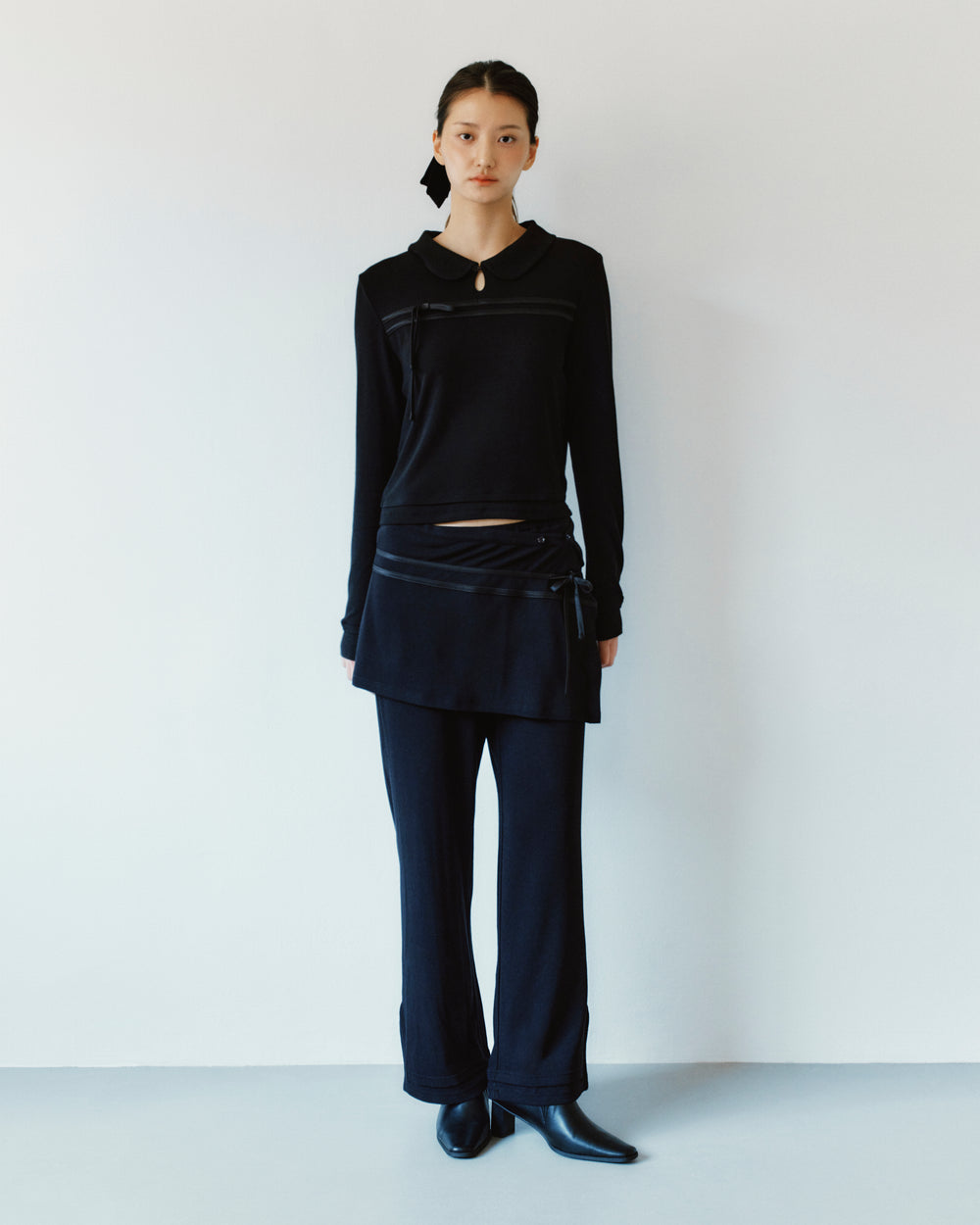 A person wearing a black long-sleeved collared top and Sundae School BLACK SEONGSU APRON PANTS, featuring wide-leg trousers with an attached hybrid wrap skirt panel.