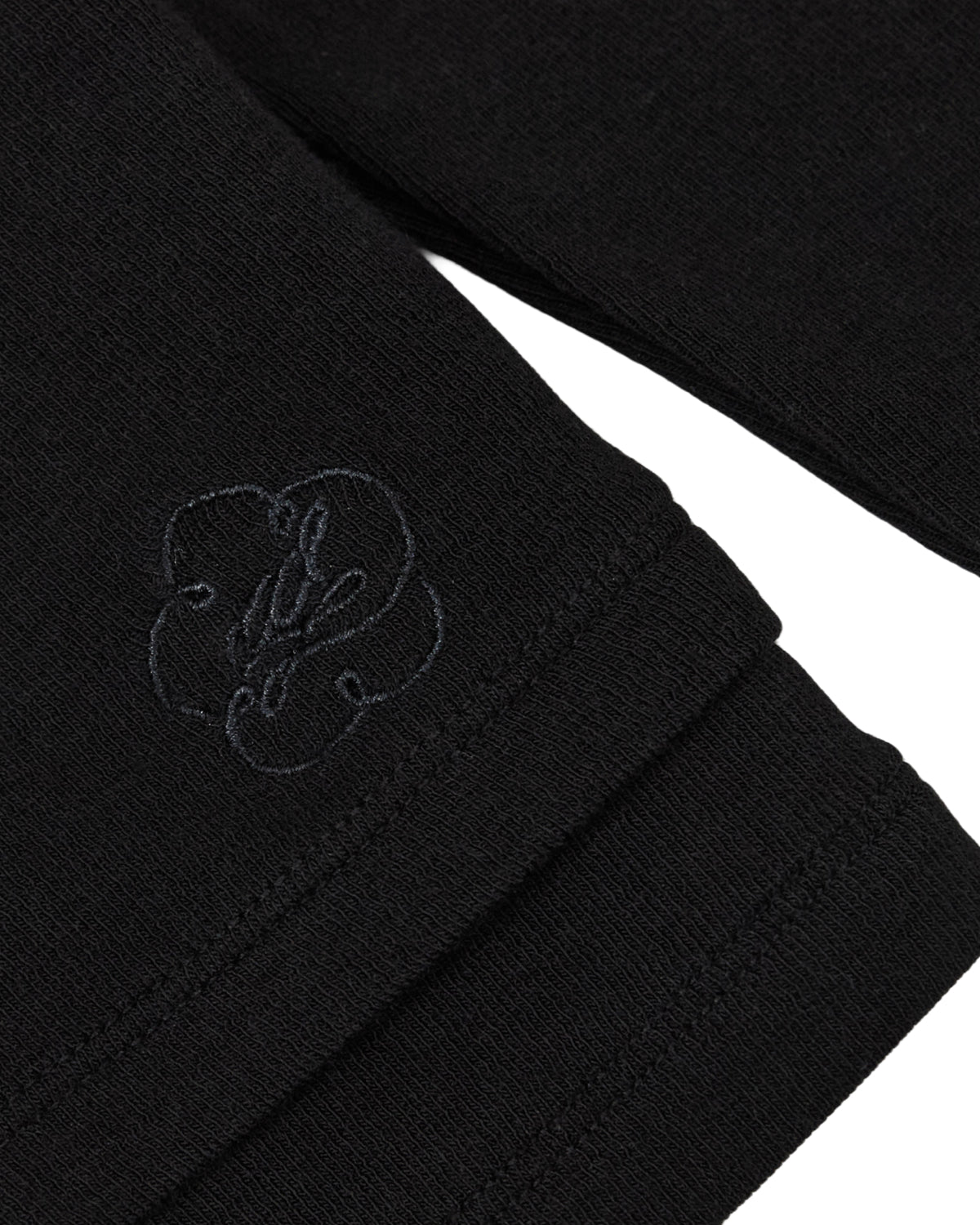 Close-up of the BLACK SEONGSU KEYHOLE TOP, a black long-sleeved blouse with a rounded collar and an embroidered floral emblem near the cuff.
