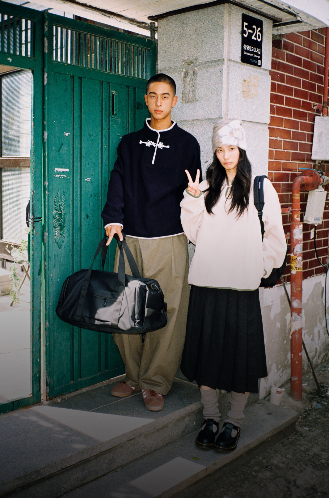 A vertical image of a young man and woman standing outdoors in casual streetwear in front of a green metal gate and brick building. The man is holding a large black duffel bag and wearing a dark blue pullover with white knot-style closures, loose khaki pants, and brown sneakers. The woman is making a peace sign and wearing a cream-colored fuzzy sweater, a fluffy white hat, a black pleated skirt, leg warmers, and black shoes.