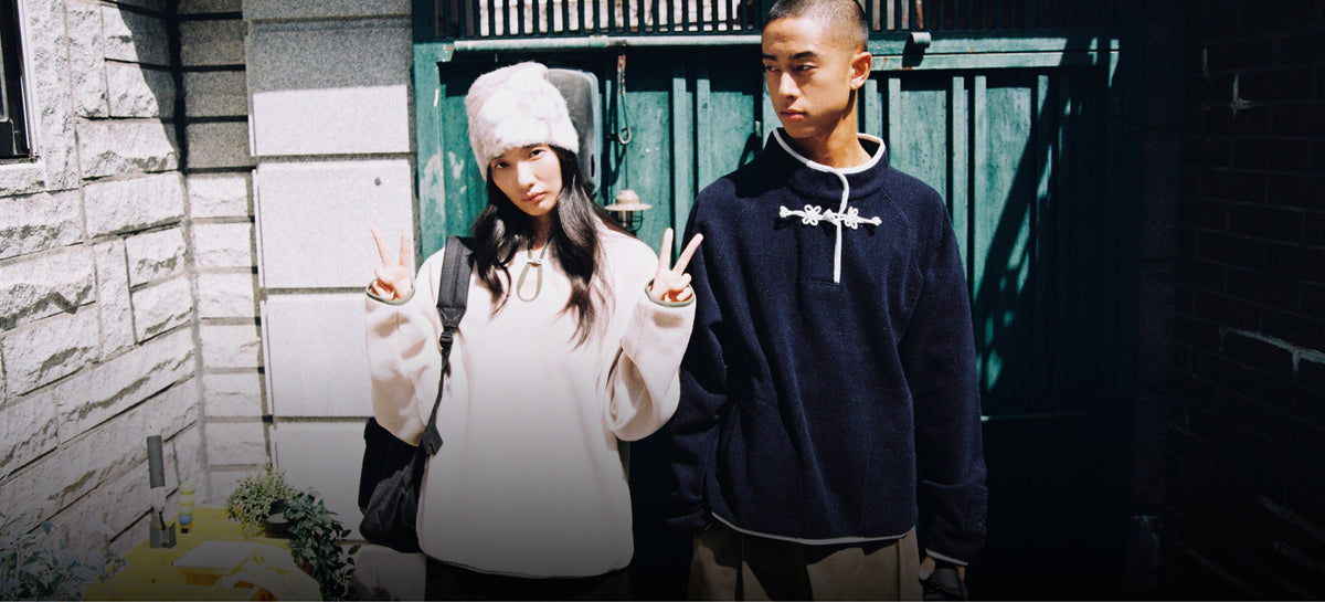 A horizontal image showing the same two models standing in bright sunlight near a stone wall and green gate. The woman stands on the left making peace signs with both hands, wearing the cream fuzzy sweater, fluffy white hat, and a large gold carabiner necklace. The man stands on the right in the dark blue pullover with white knot closures, looking off-camera to the side.