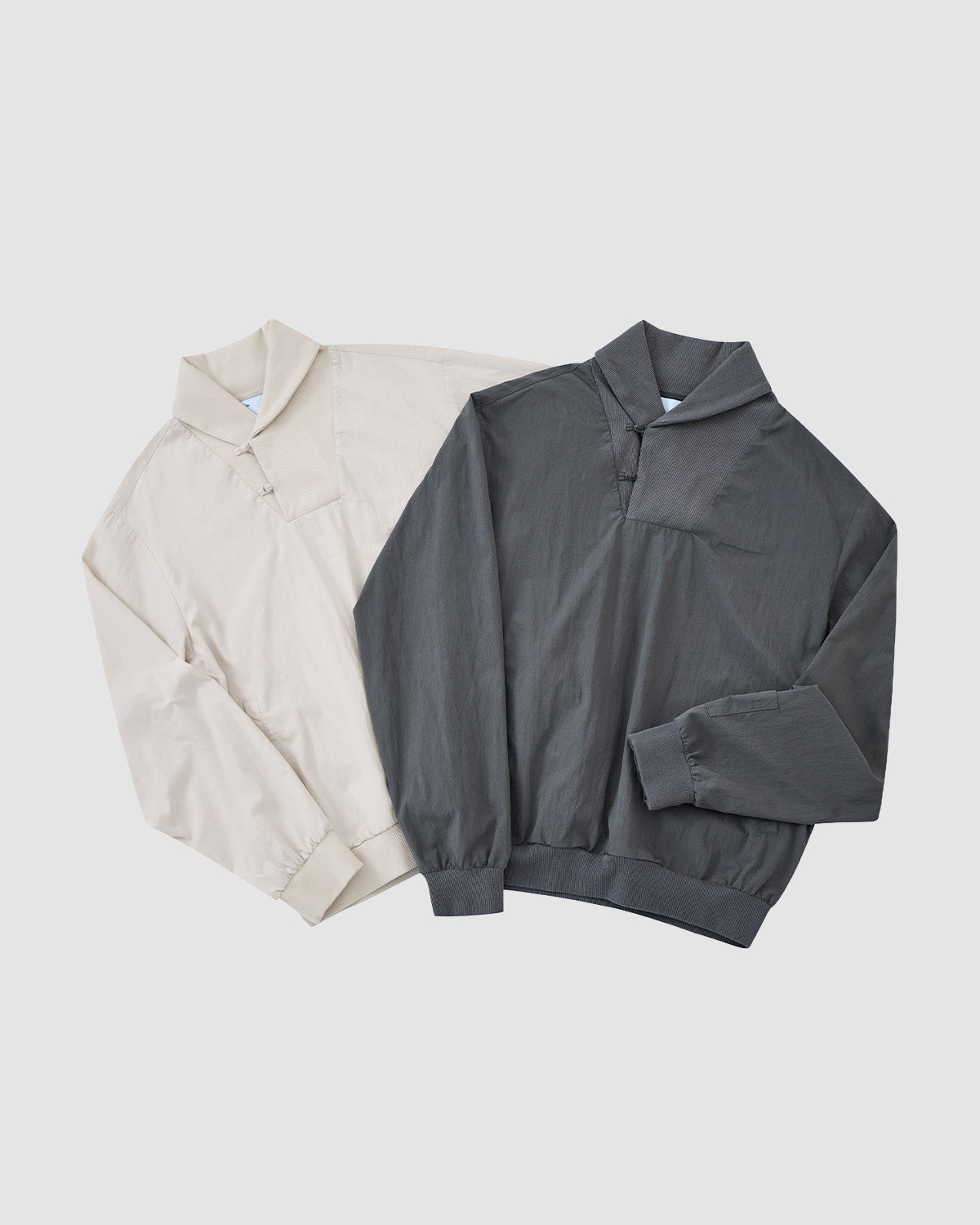 Two Sundae School CHARCOAL SHAWL COLLAR ANORAK jackets, in beige and dark gray, are laid flat side by side. Each features a shawl collar, front zipper, and elastic cuffs and waistband for a subtle monk-core vibe.