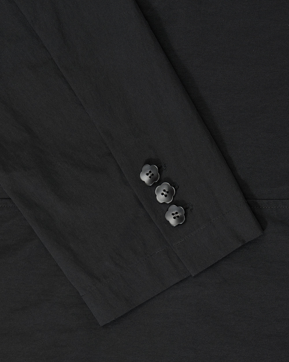 Close-up of the BLACK STAND COLLAR UNIFORM JACKET by Sundae School, showing its black sleeve with three black buttons near the cuff, crafted from a cotton nylon blend and set against matching fabric.