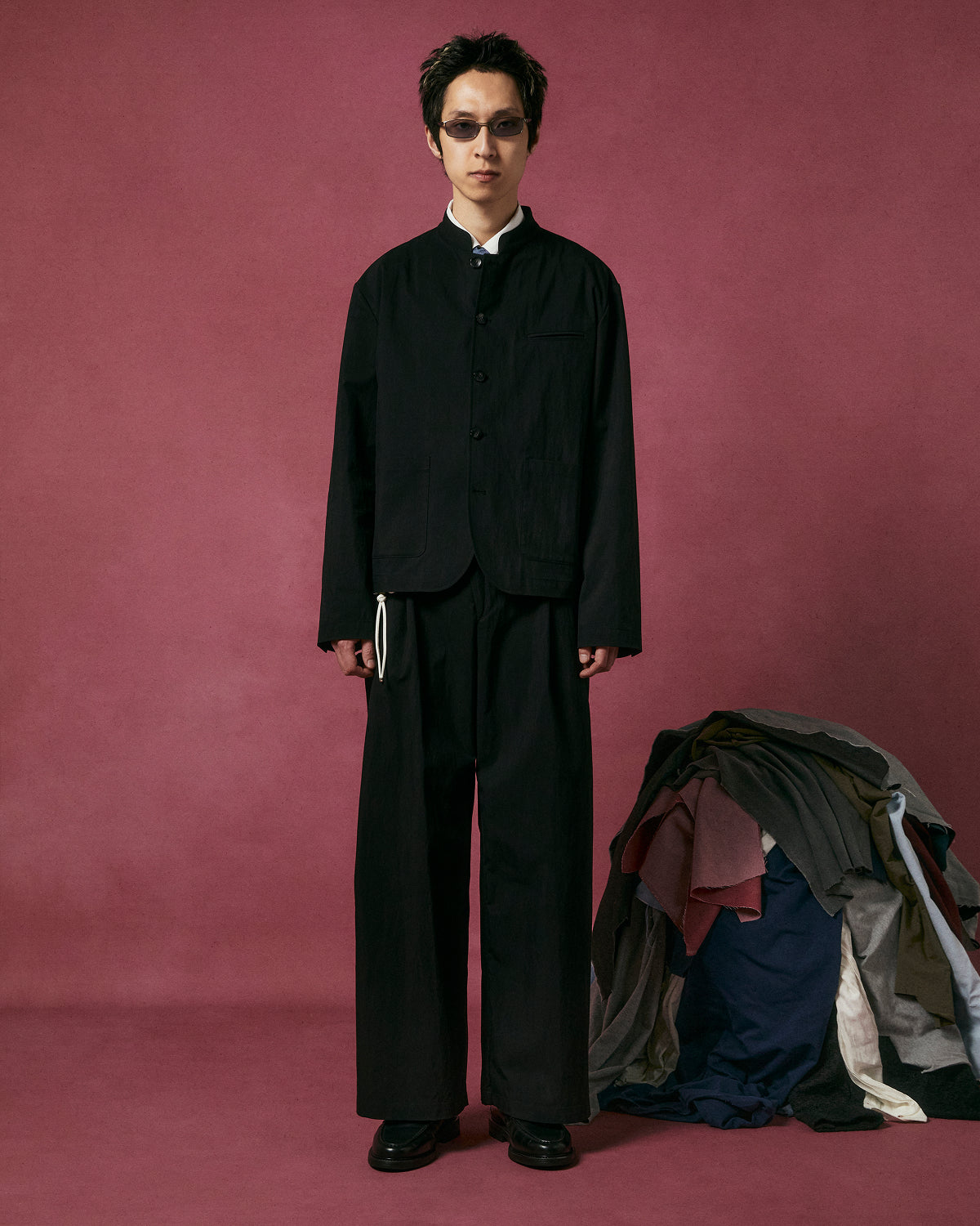 A person in Sundae School BLACK PLEATED UNIFORM SLACKS and black sunglasses stands before a pink backdrop, beside a pile of assorted clothes stacked on the floor.