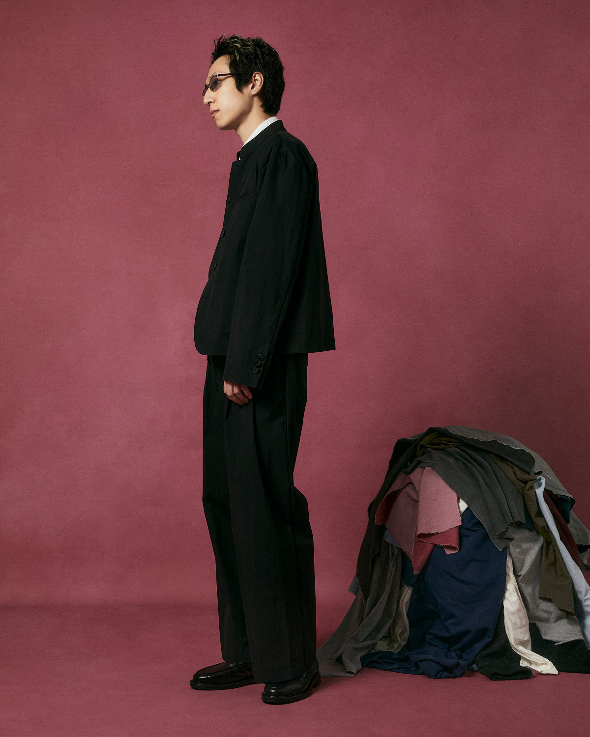 A man in a black suit and sunglasses stands in profile against a maroon background next to a pile of clothing, featuring Sundae School BLACK PLEATED UNIFORM SLACKS.