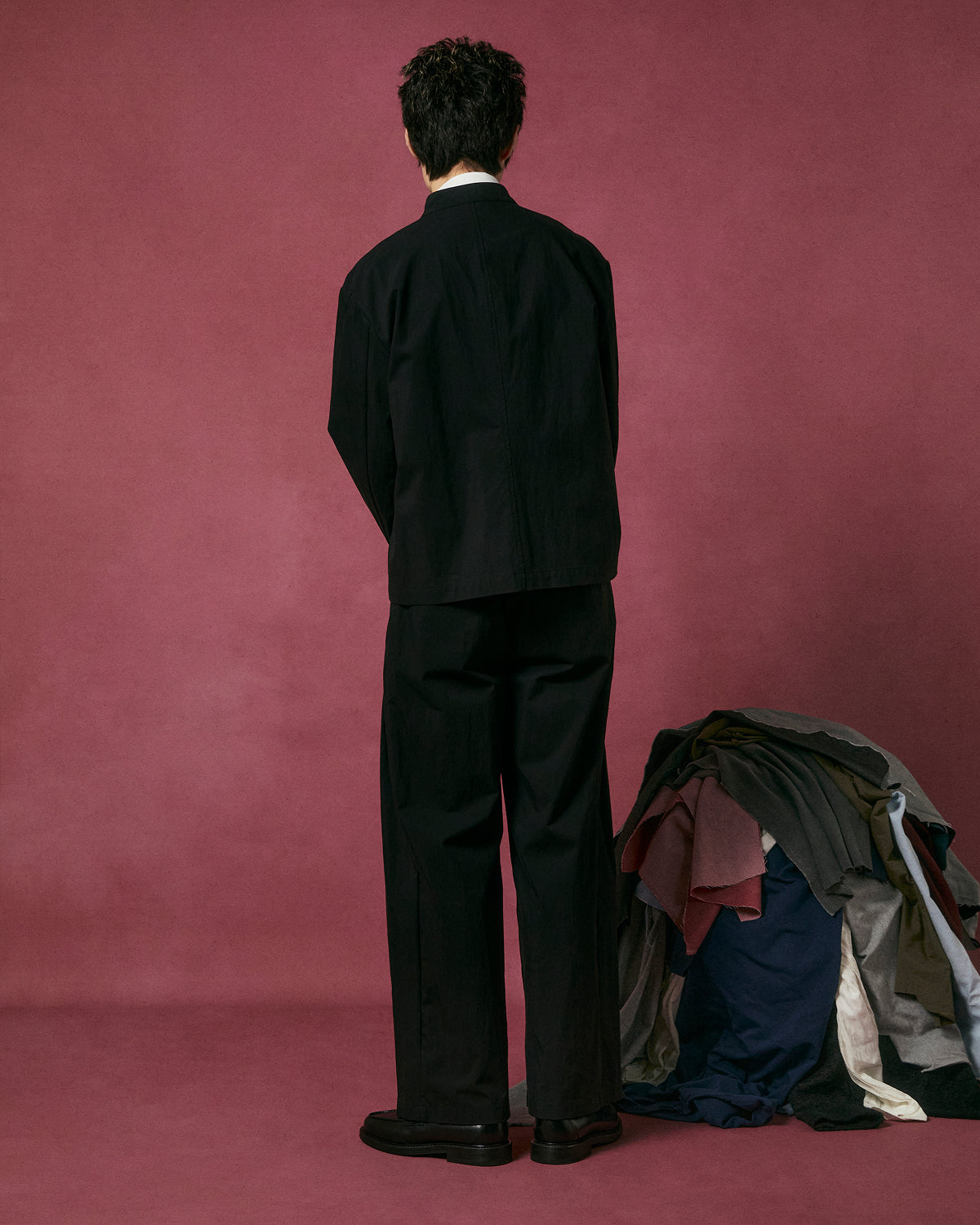 A person in Sundae School BLACK PLEATED UNIFORM SLACKS stands with their back to the camera against a maroon background. To their right, assorted folded clothes in various colors are stacked on the floor.