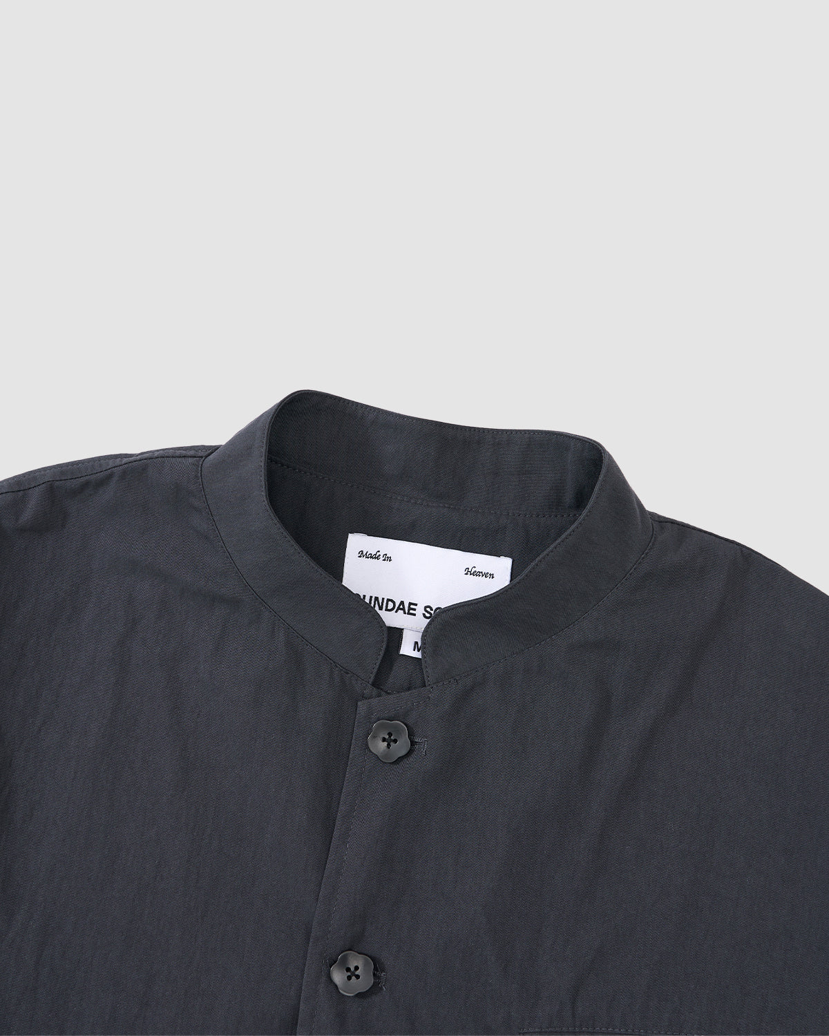 A close-up of the Sundae School CHARCOAL STAND COLLAR UNIFORM JACKET, made from a soft cotton-nylon blend with two buttons, set against a light grey background. A white label is visible inside the collar.