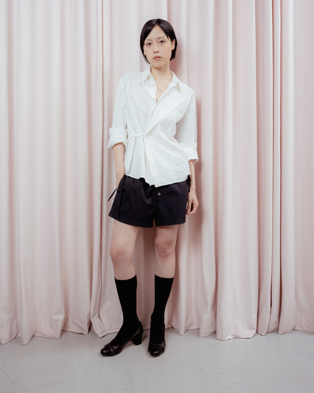A person with short dark hair stands in front of light pink curtains, wearing a white button-up shirt, Sundae School STRIPED BOXER SHORTS, long black socks, and black shoes.