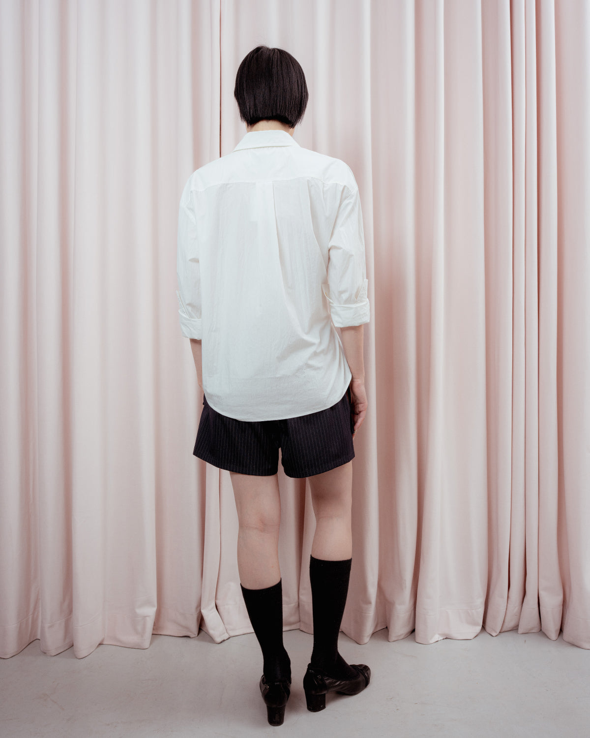 Full-length back view of a person with short black hair standing against a light pink curtain backdrop. They are wearing a loose white button-down shirt, dark pinstripe boxer-style shorts, mid-calf black socks, and black low-heeled shoes.