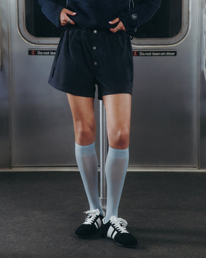A person stands on a subway train wearing a navy hoodie, Sundae School NAVY STRIPE BOXER SHORTS, pale blue knee-high socks, and black-and-white sneakers. Their hands are in their pockets with subway doors in the background.
