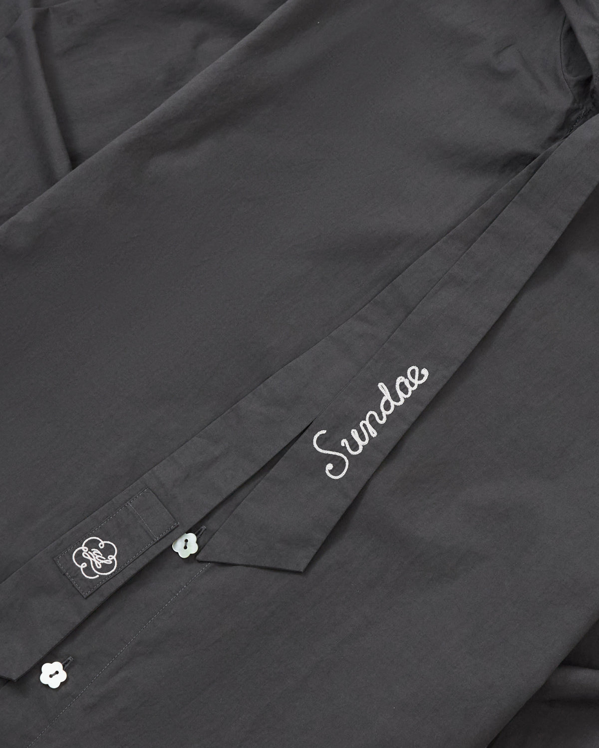 A close-up of the CHARCOAL SUNBAE TIE SHIRT by Sundae School shows white embroidery—one area spells out “Sundae,” while another features a small flower-shaped button topped with a cloud design.