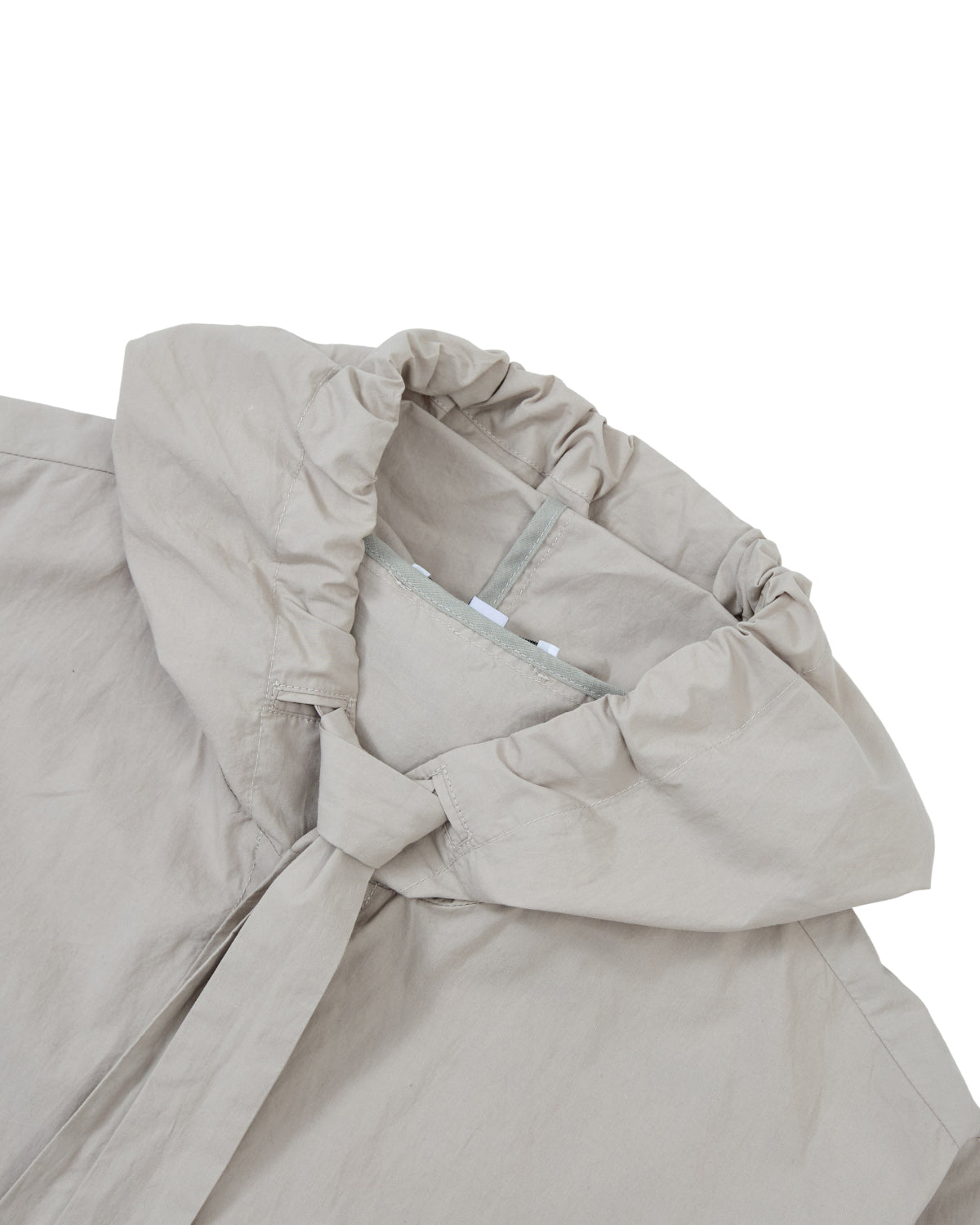 A close-up of the GREY SUNBAE TIE SHIRT by Sundae School, featuring a light beige fabric with a gathered, elastic hood and a partially visible collar, laid flat on a white surface.