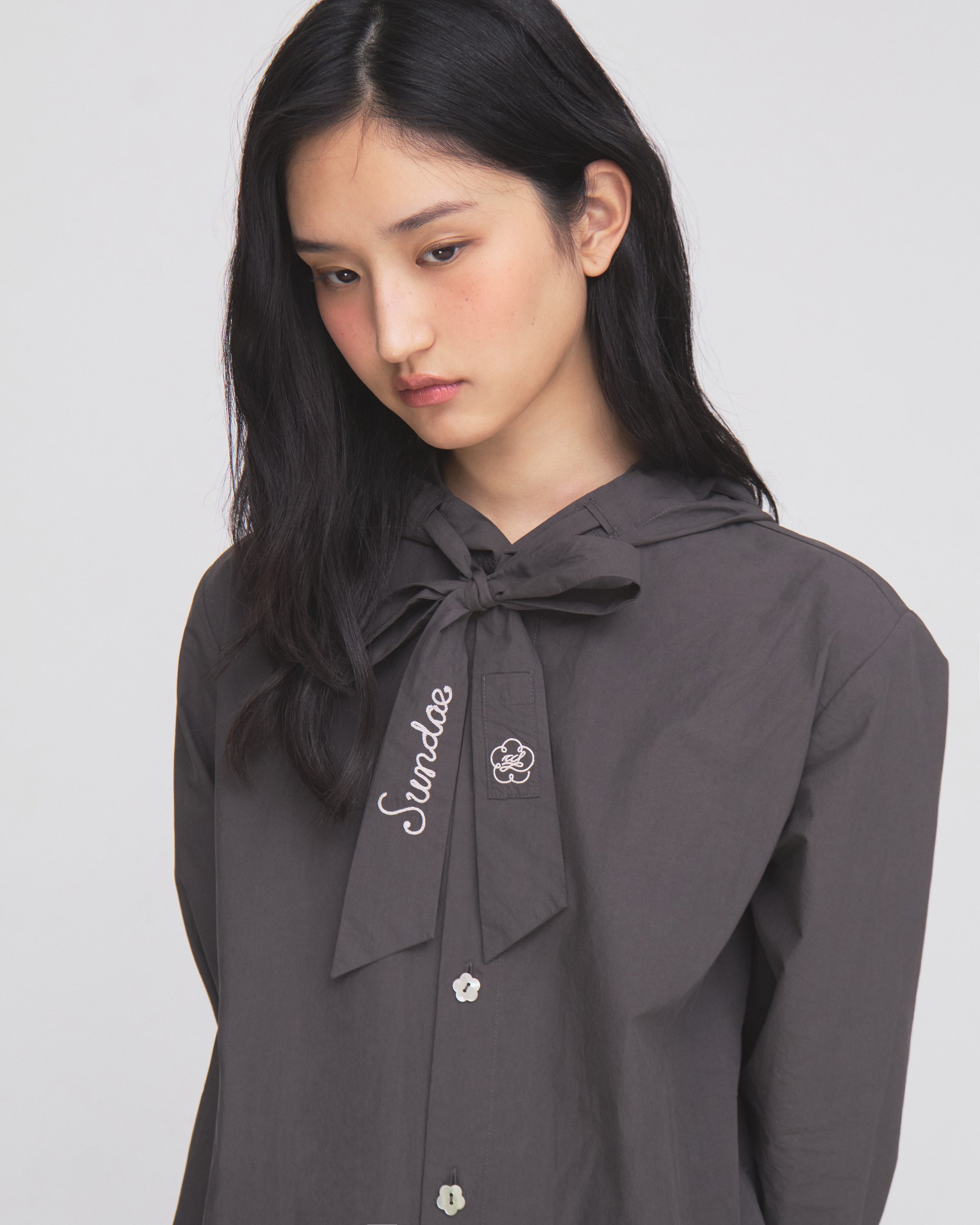 A woman with long black hair wears the CHARCOAL SUNBAE TIE SHIRT by Sundae School, featuring a dark gray button-up design and a large bow at the collar embroidered with “Sunday.” She looks down with a neutral expression against a plain background.