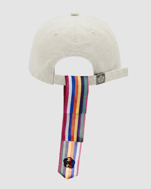 The Sundae School IVORY DAENGGI CAP is a light-colored adjustable cotton cap seen from the back, with a rainbow-striped daenggi attached to the strap and hanging below, featuring a flower patch at the end for a playful accent.