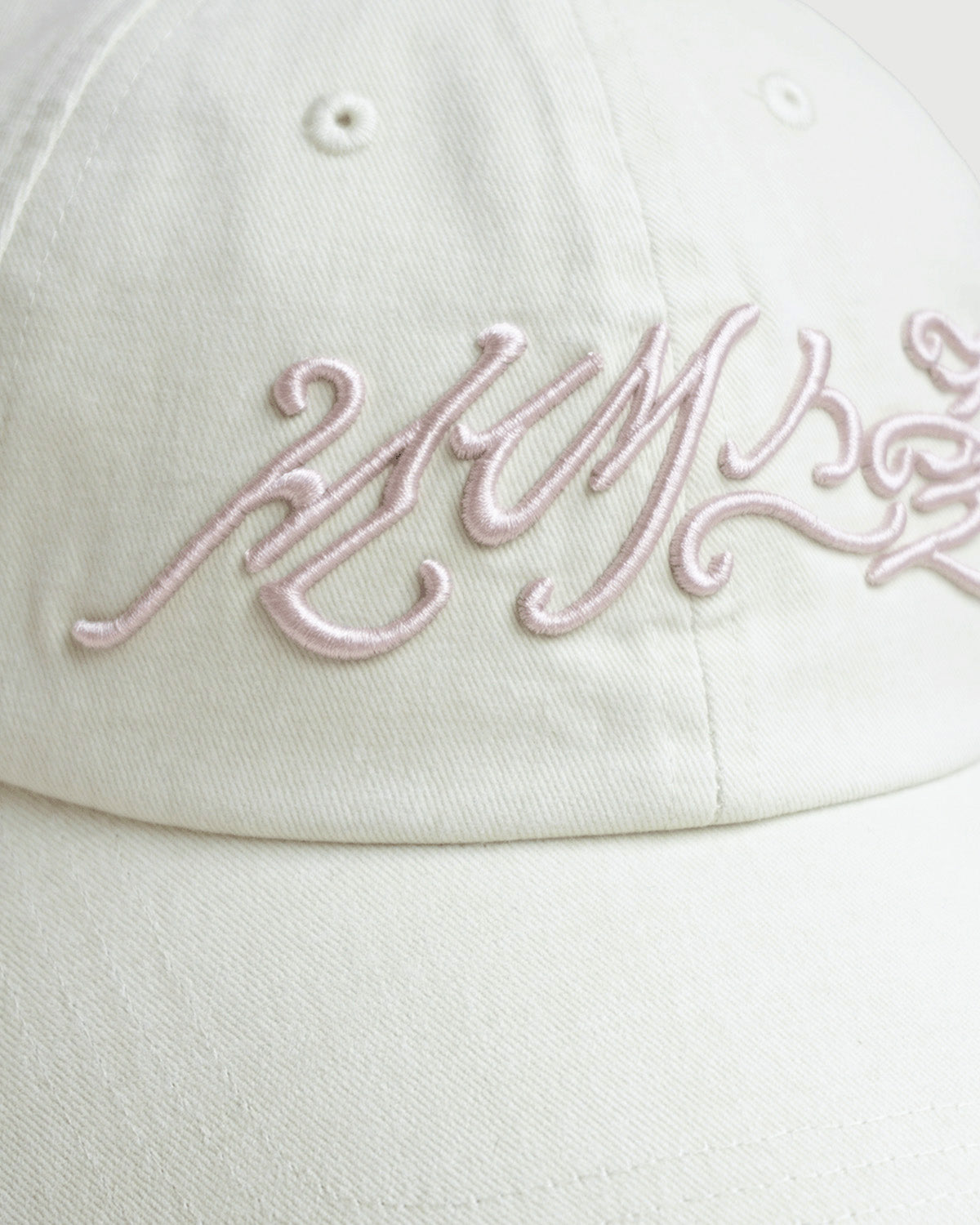 A close-up of the IVORY DAENGGI CAP by Sundae School, an adjustable cotton hat in white, featuring intricate pale pink embroidery in a stylized script across the front.