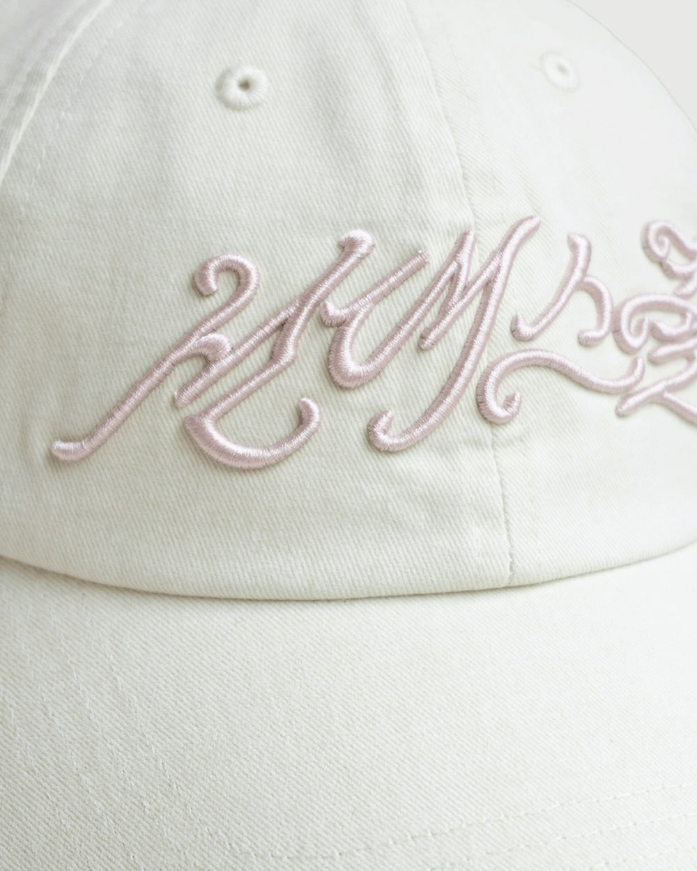 A close-up of the IVORY DAENGGI CAP by Sundae School, an adjustable cotton hat in white, featuring intricate pale pink embroidery in a stylized script across the front.
