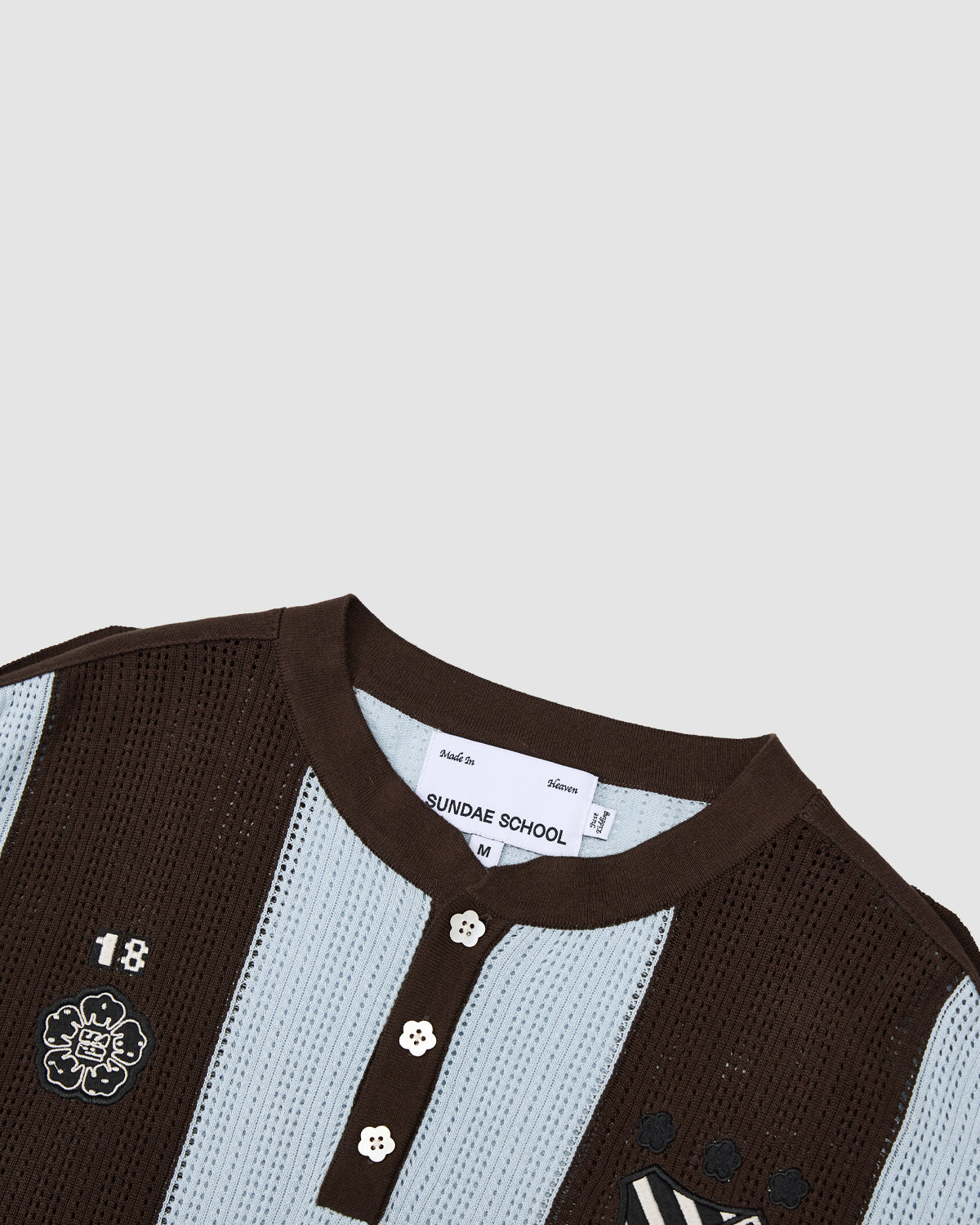 A close-up of the SKY BLUE SUNDAE SOCCER JERSEY by Sundae School shows a lightweight blue and brown knit shirt with a round collar, three floral mochi buttons, embroidered chest patches, and an inner neck brand tag.