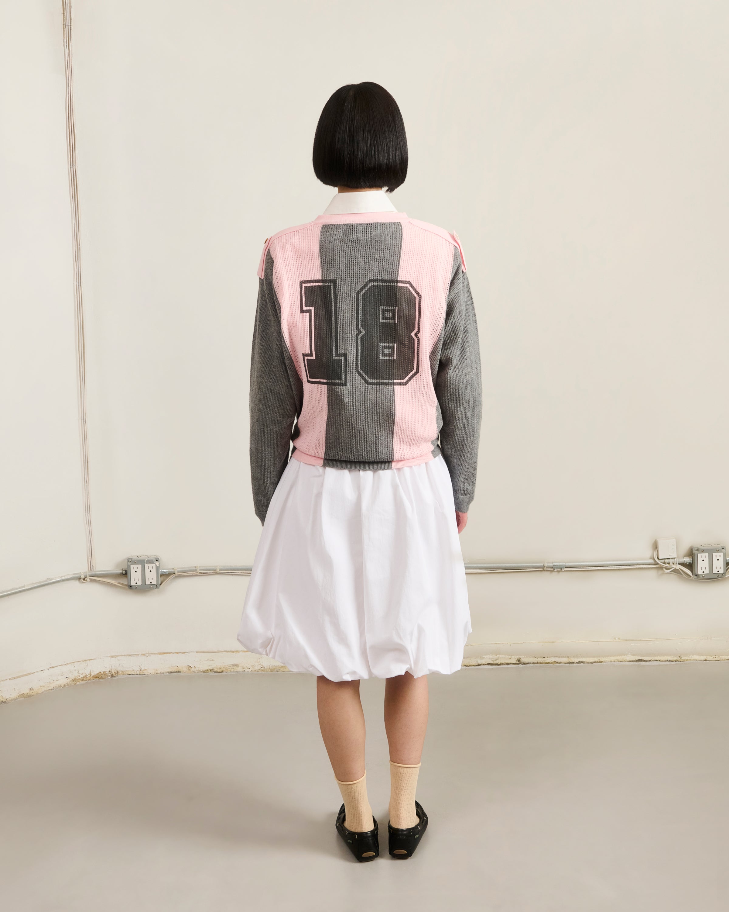 A person with a short black bob stands facing a plain beige wall, wearing Sundae School’s PINK SUNDAE SOCCER JERSEY with embroidered patches and the number 18 on the back, paired with a white skirt, beige socks, and black shoes.