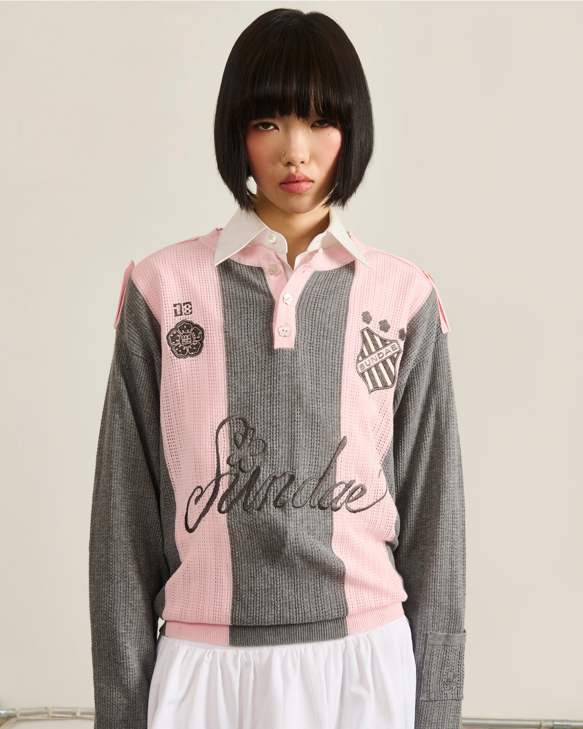 A young woman with a straight black bob wears the Sundae School PINK SUNDAE SOCCER JERSEY—a gray and pink, 100% cotton top with custom embroidery—paired with a white pleated skirt, standing against a plain background.