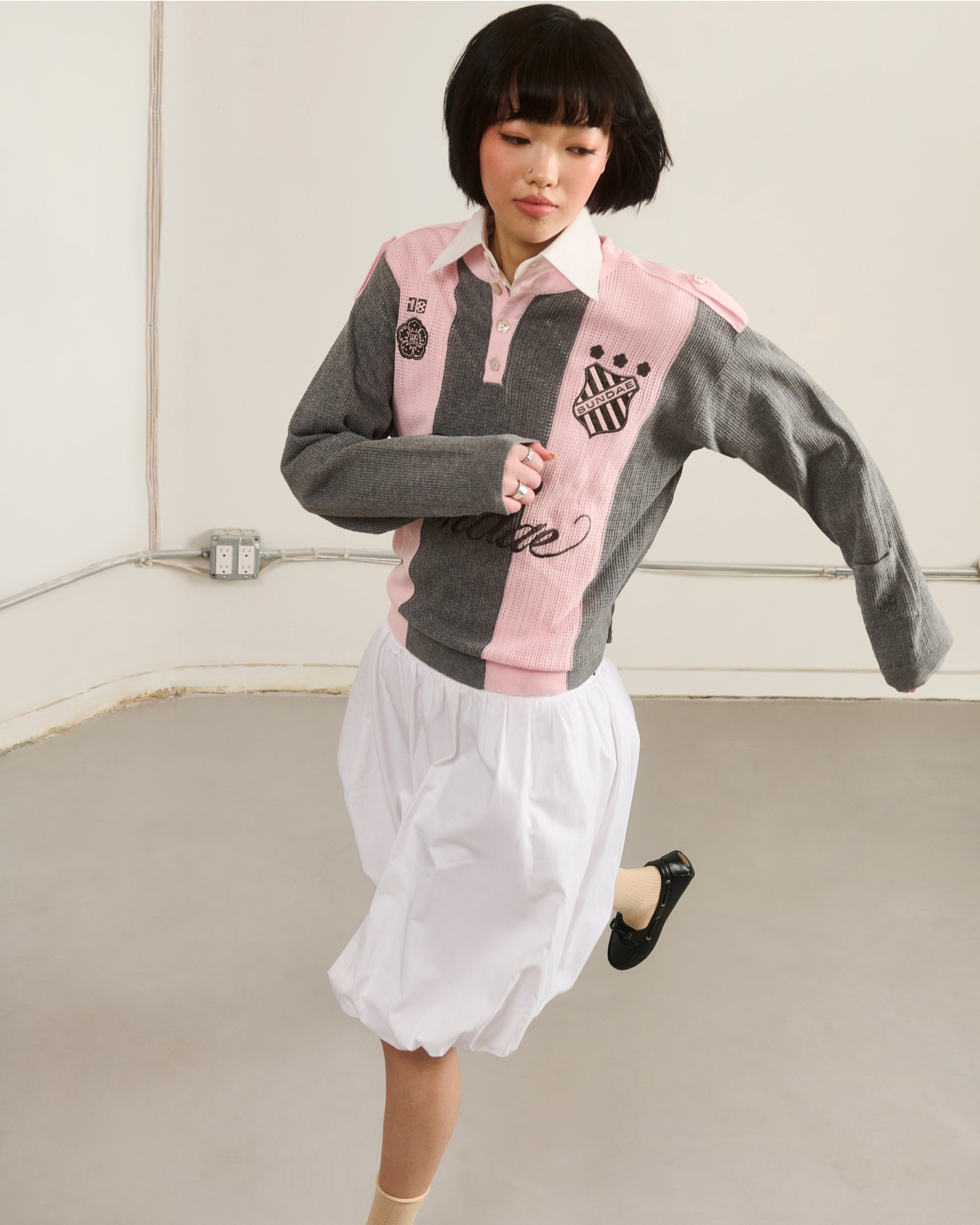 A young woman with short dark hair moves in a minimal room, wearing Sundae School’s PINK SUNDAE SOCCER JERSEY—a 100% cotton lightweight top in pink and gray—over a white skirt and black shoes, set against bare walls with exposed outlets.