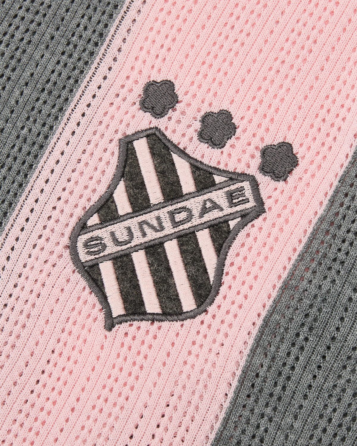 A close-up of the PINK SUNDAE SOCCER JERSEY by Sundae School highlights its pink and grey striped perforated knit fabric, featuring a black and white shield patch with “SUNDAE” text and three small paw prints above it. This lightweight top is crafted from 100% cotton and finished with custom embroidered chest patches for a unique touch.