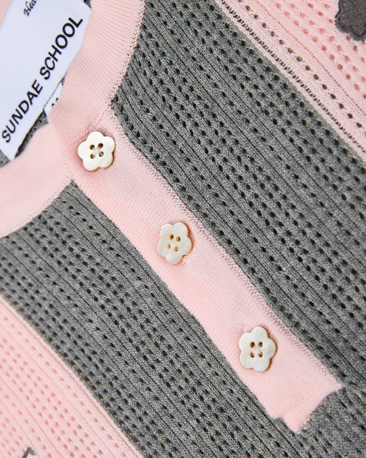 Close-up of the Sundae School PINK SUNDAE SOCCER JERSEY, a perforated knit garment in pink and gray, detailed with three small white flower-shaped buttons and a visible Sundae School label.