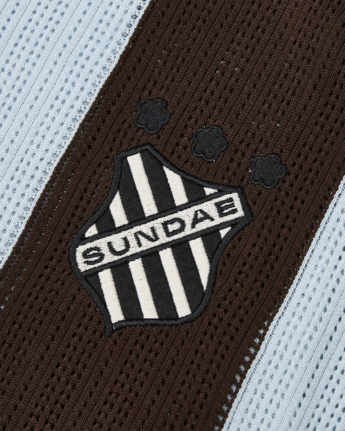 A black and white striped shield patch featuring the word “SUNDAE” and three small flower motifs—evoking floral mochi buttons—is sewn onto a brown and sky blue textured fabric, adding charm to this cotton lightweight SKY BLUE SUNDAE SOCCER JERSEY from Sundae School.