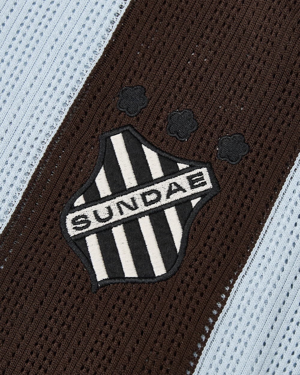 A black and white striped shield patch featuring the word “SUNDAE” and three small flower motifs—evoking floral mochi buttons—is sewn onto a brown and sky blue textured fabric, adding charm to this cotton lightweight SKY BLUE SUNDAE SOCCER JERSEY from Sundae School.