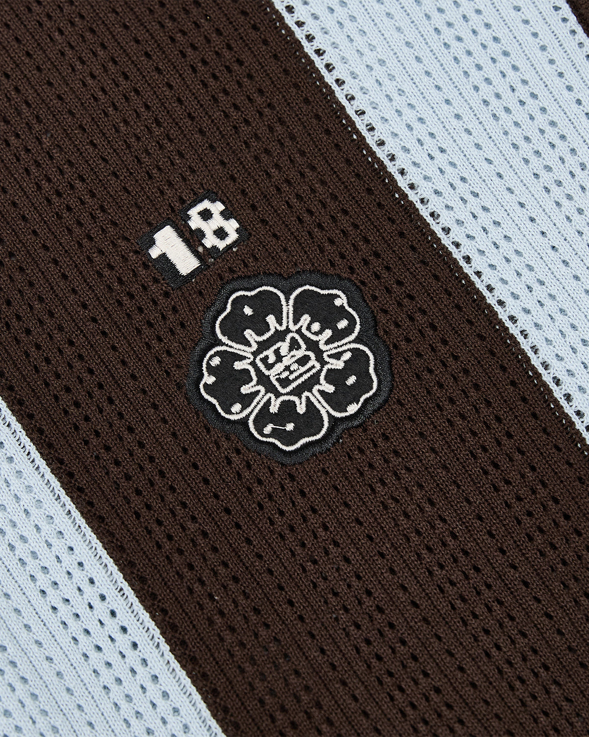 Close-up of the SKY BLUE SUNDAE SOCCER JERSEY by Sundae School, a cotton lightweight top crafted from knit fabric with vertical black and light blue stripes, featuring a white number 18 and an embroidered flower emblem with stylized faces in each petal.