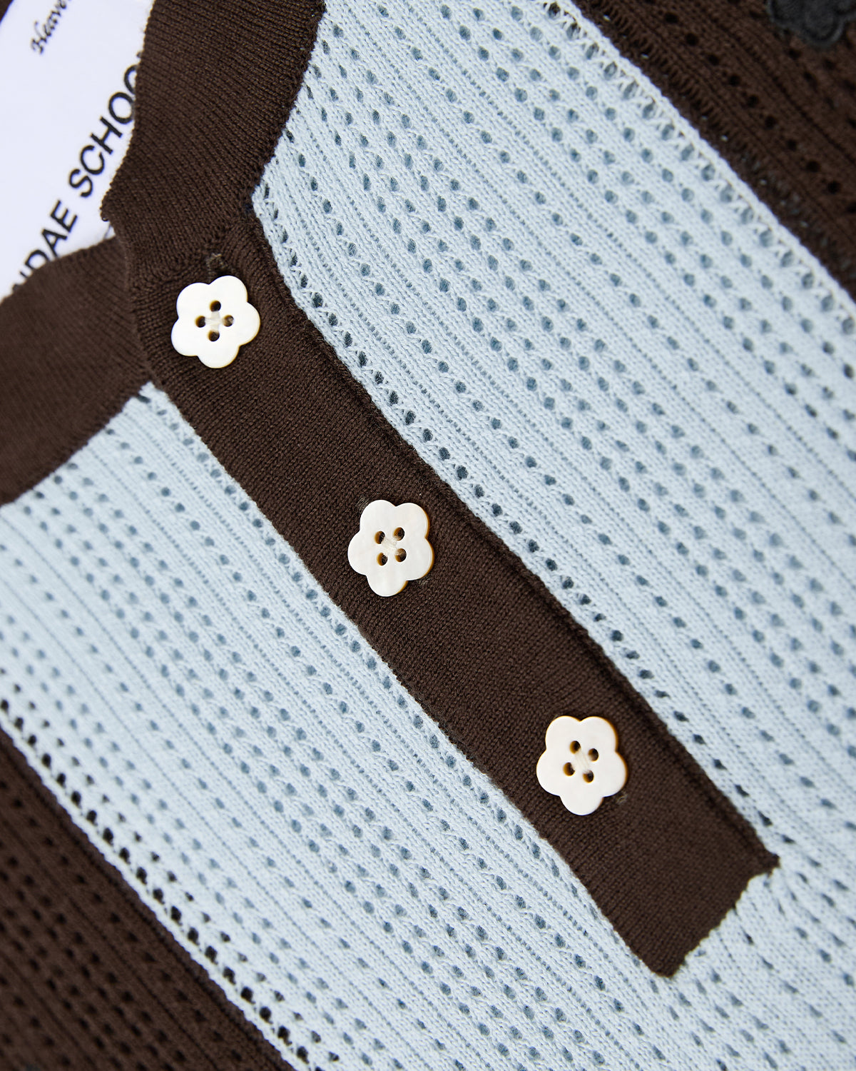 A close-up of the textured SKY BLUE SUNDAE SOCCER JERSEY by Sundae School, featuring a blue and brown knit with three white floral mochi buttons at the center. Part of a white tag is partially visible in the top left corner.