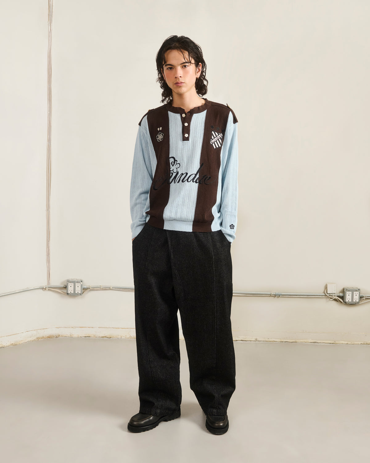 A person with long dark hair stands indoors against a light wall, wearing the Sundae School SKY BLUE SUNDAE SOCCER JERSEY with wide-leg dark pants and black shoes; electrical outlets and cords are visible.