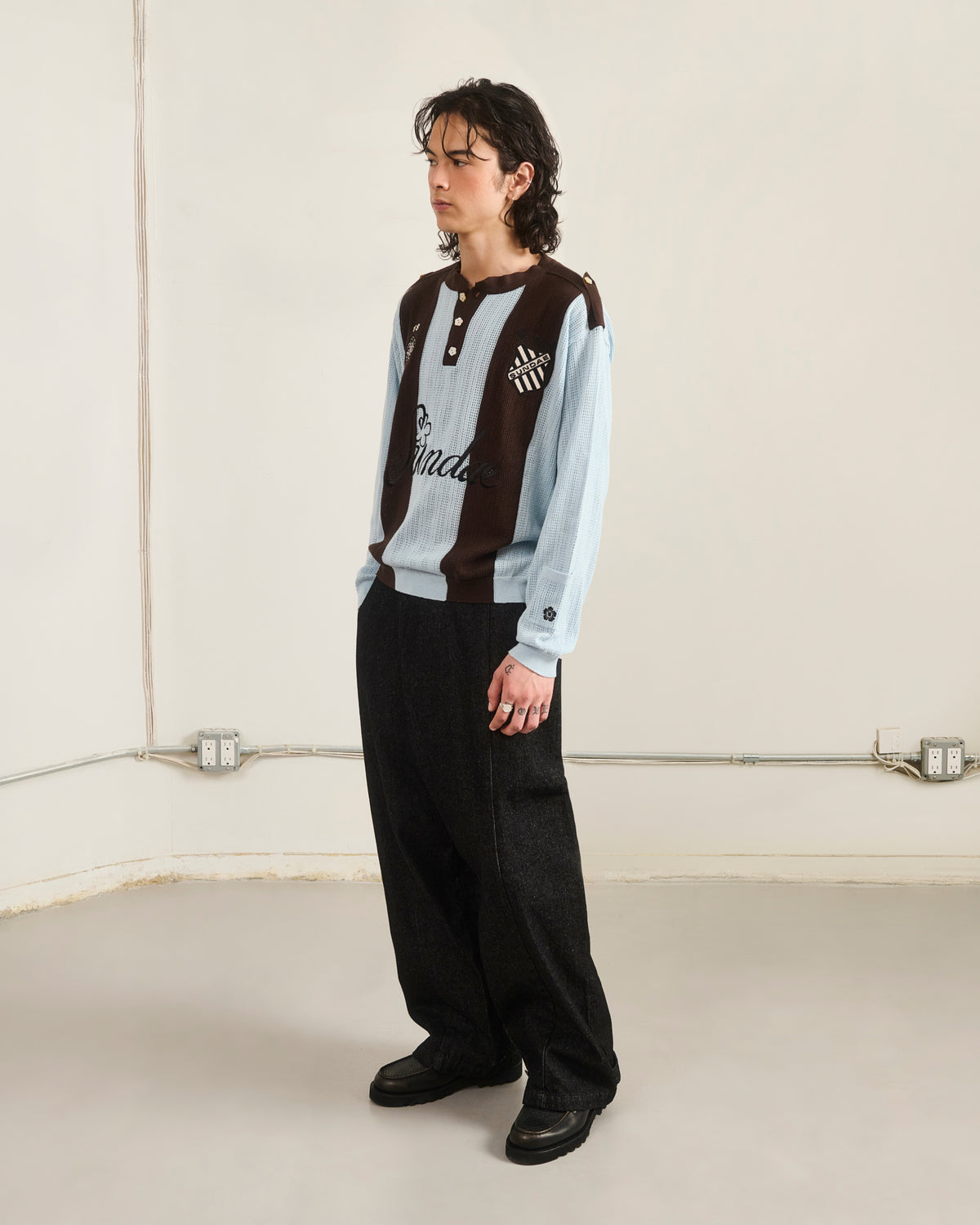 A person with shoulder-length dark hair stands in a minimal room wearing the Sundae School SKY BLUE SUNDAE SOCCER JERSEY, wide black pants, and black shoes. The beige walls have visible outlets and baseboards.