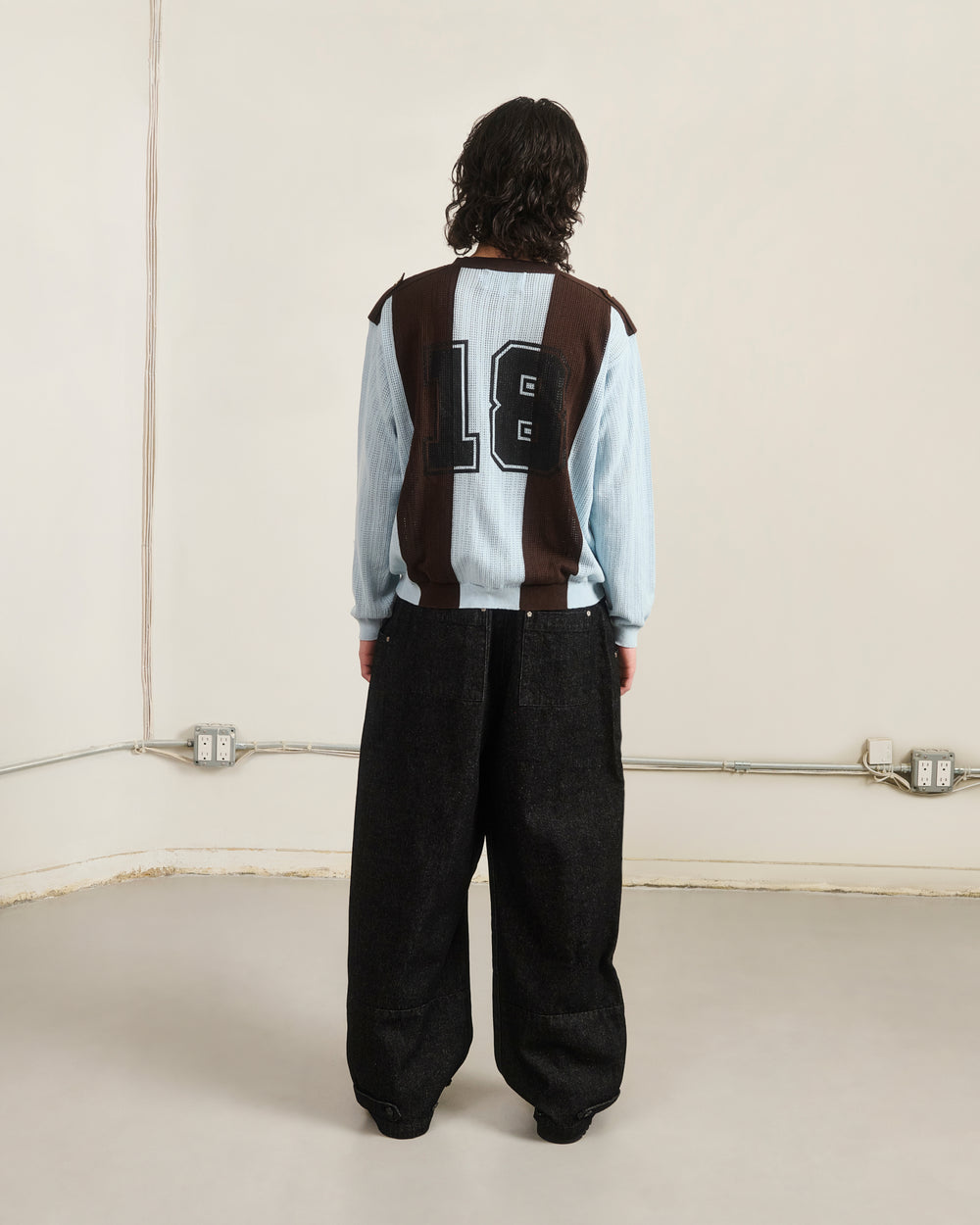 Facing a plain wall with visible outlets and cords, a person with shoulder-length hair wears the Sundae School SKY BLUE SUNDAE SOCCER JERSEY, featuring the number 18 and floral mochi buttons, paired with loose black pants.