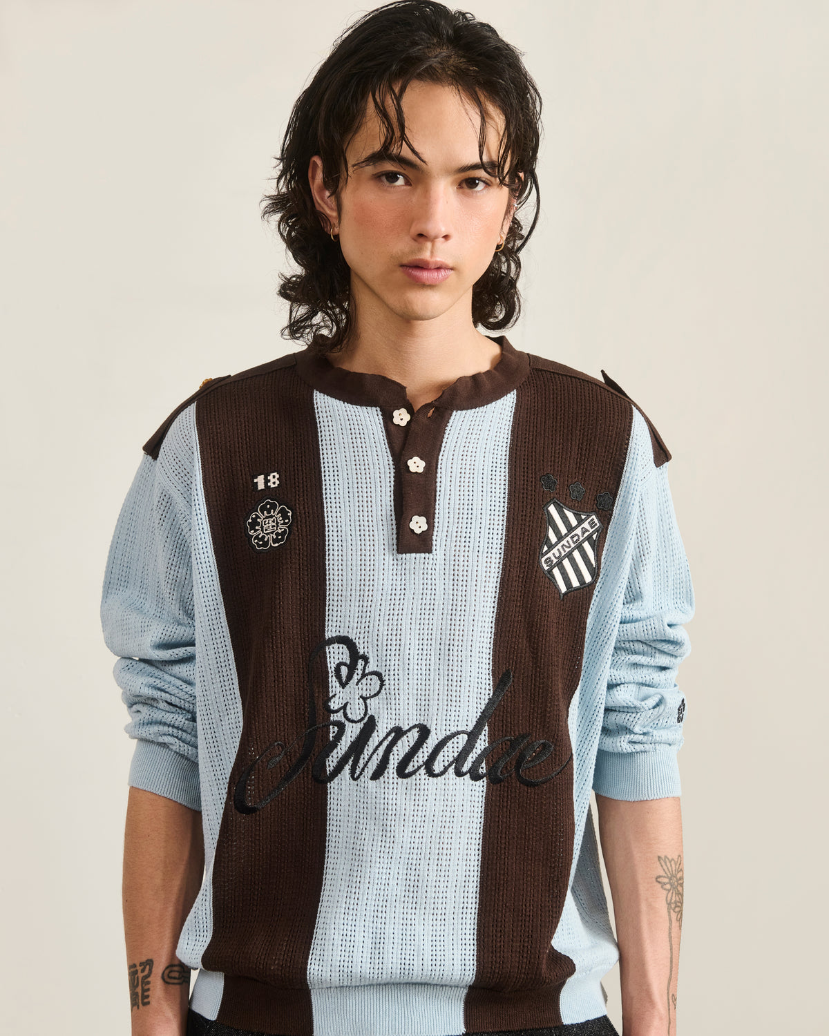 A person with wavy dark hair wears the SKY BLUE SUNDAE SOCCER JERSEY by Sundae School, featuring light blue and brown stripes, floral mochi buttons, and embroidered chest patches, standing against a plain background.