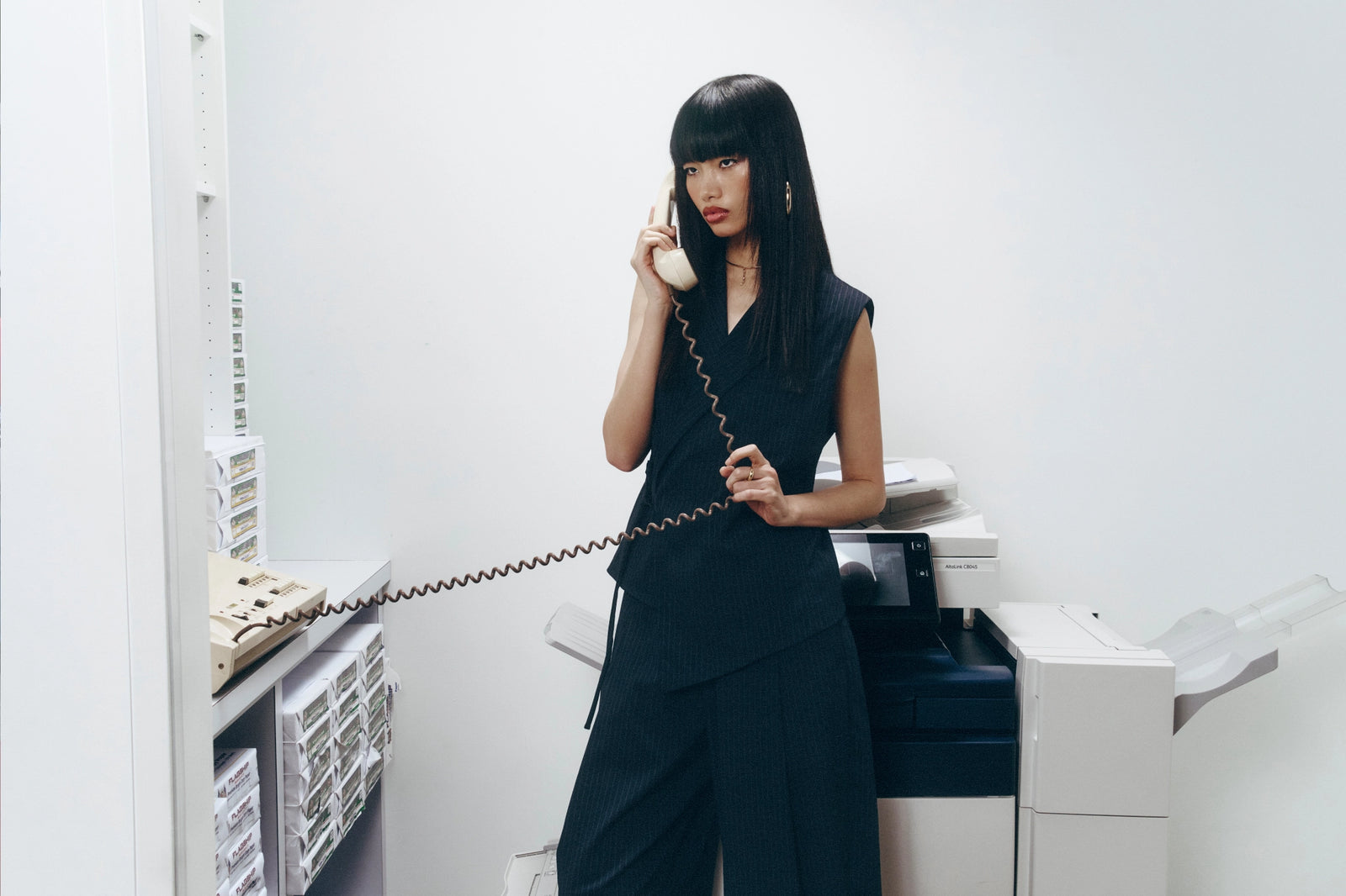 A woman with long black hair stands in a minimalist office setting, holding a vintage cream rotary telephone receiver to her ear. She is wearing a dark navy sleeveless top and matching wide-leg trousers.