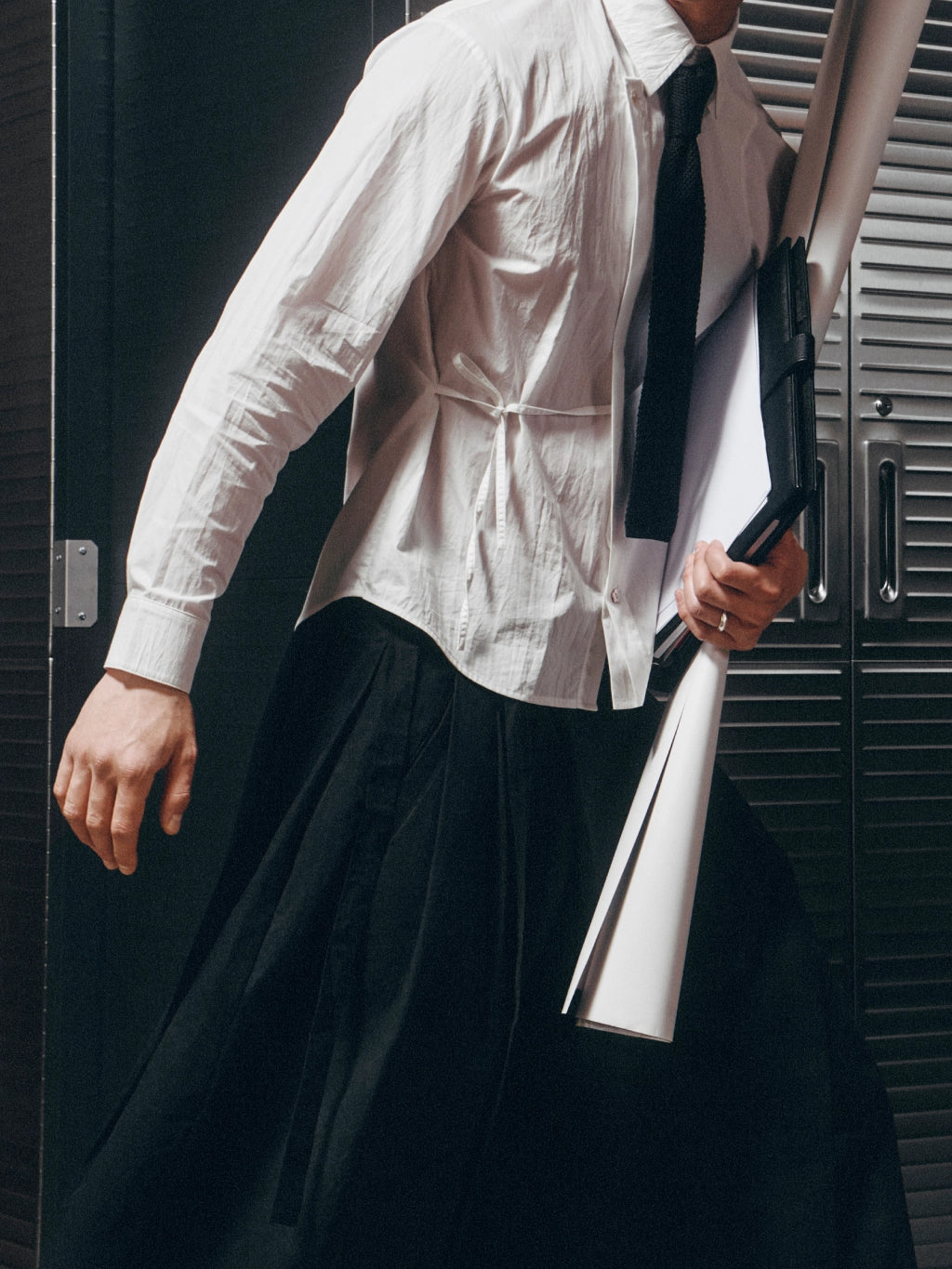A person wearing a white long-sleeve button-up shirt and black tie, holding rolled white sheets or a folder in one hand, standing in front of dark metal lockers.