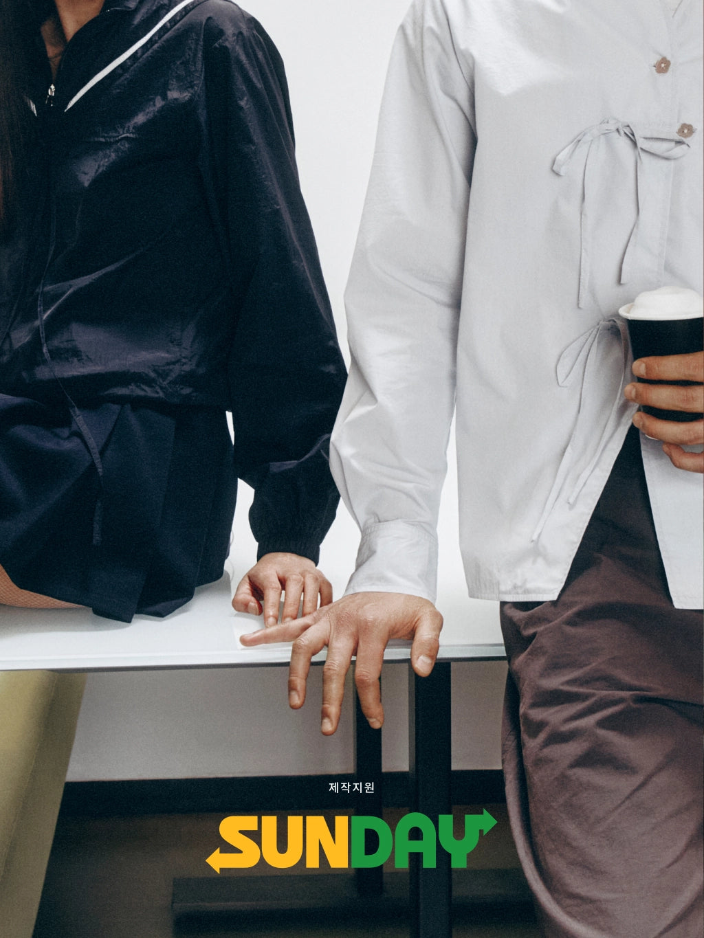A fashion editorial-style photo showing two people seated at a white table, cropped from the shoulders down. The person on the left wears a shiny black jacket with white piping, and the person on the right wears a loose white long-sleeve top and brown pants while holding a takeaway coffee cup.