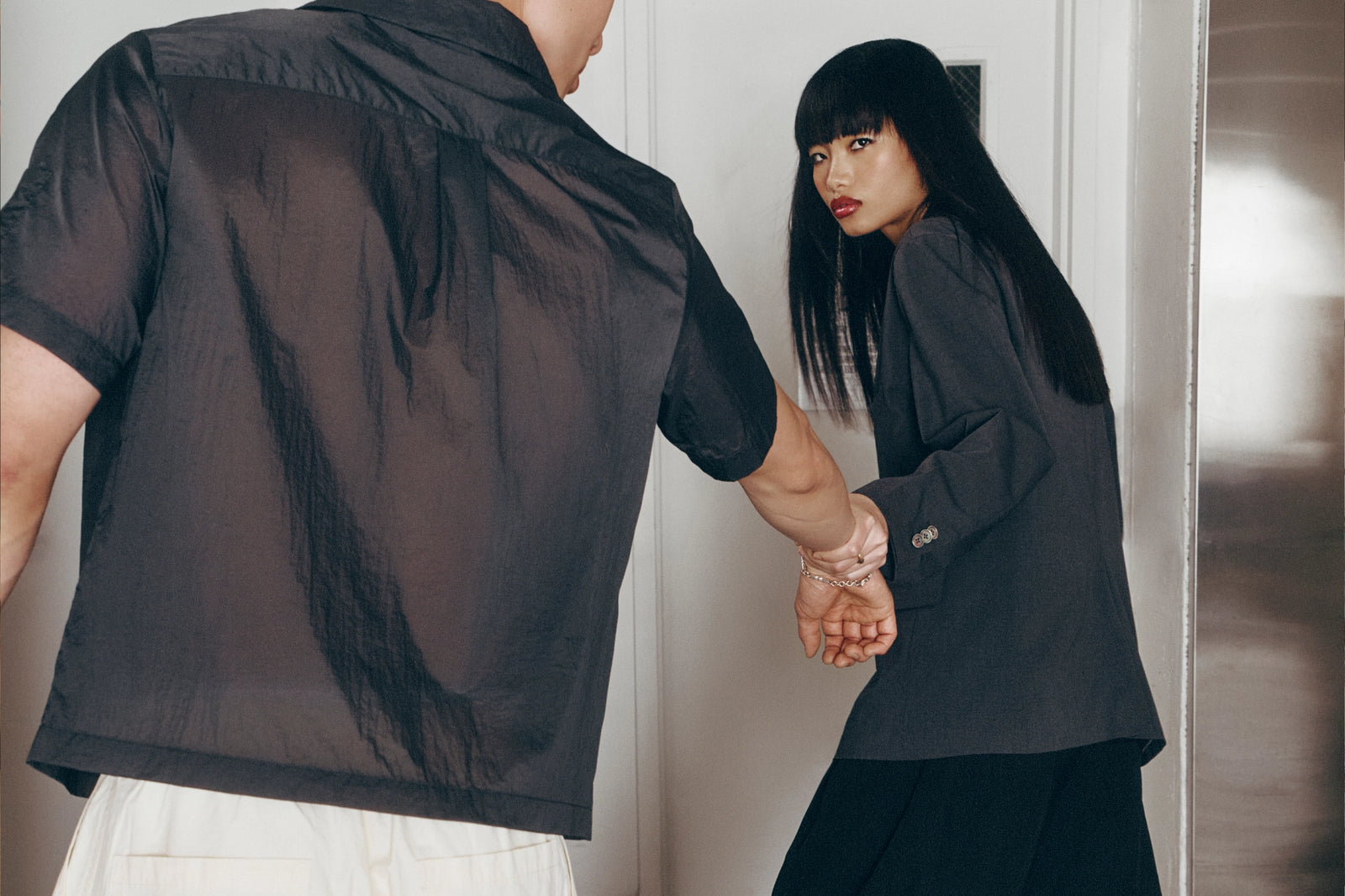 A person shown from behind wearing a sheer black short-sleeve shirt, holding hands with a woman wearing a dark gray blazer and black skirt in an indoor setting.