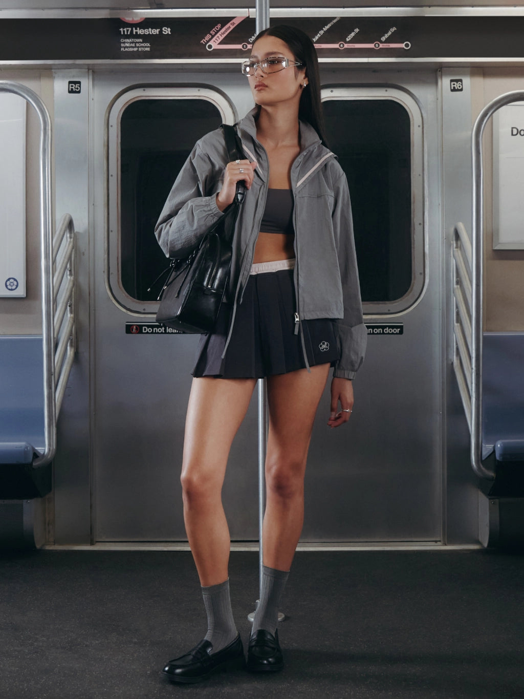 A woman stands on a subway train holding a pole, wearing a gray zip-up jacket, black athletic crop top and shorts, gray socks, black loafers, and carrying a black shoulder bag.
