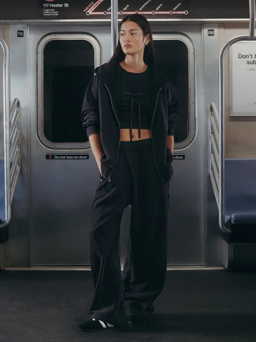A model wearing a black streetwear outfit consisting of a zip-up hoodie, cropped tie-front top, and wide-leg pants, standing inside a subway train car.