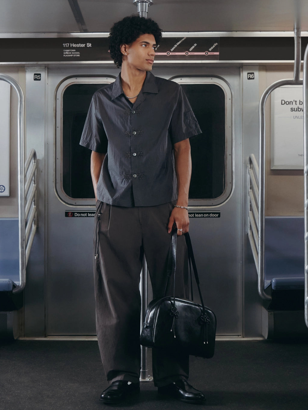 A man stands on a subway train wearing a dark gray short-sleeve button-up shirt and matching dark gray trousers, holding a black duffel bag.